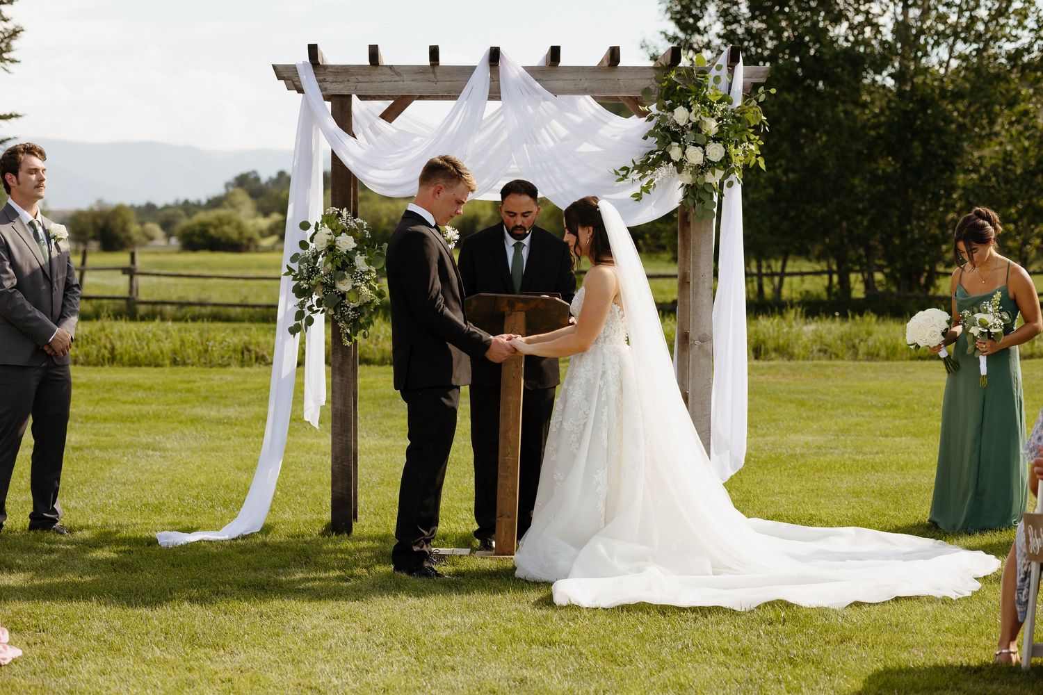 Outdoor wedding ceremony under wooden pergola with white draping as couple exchanges vows at altar.