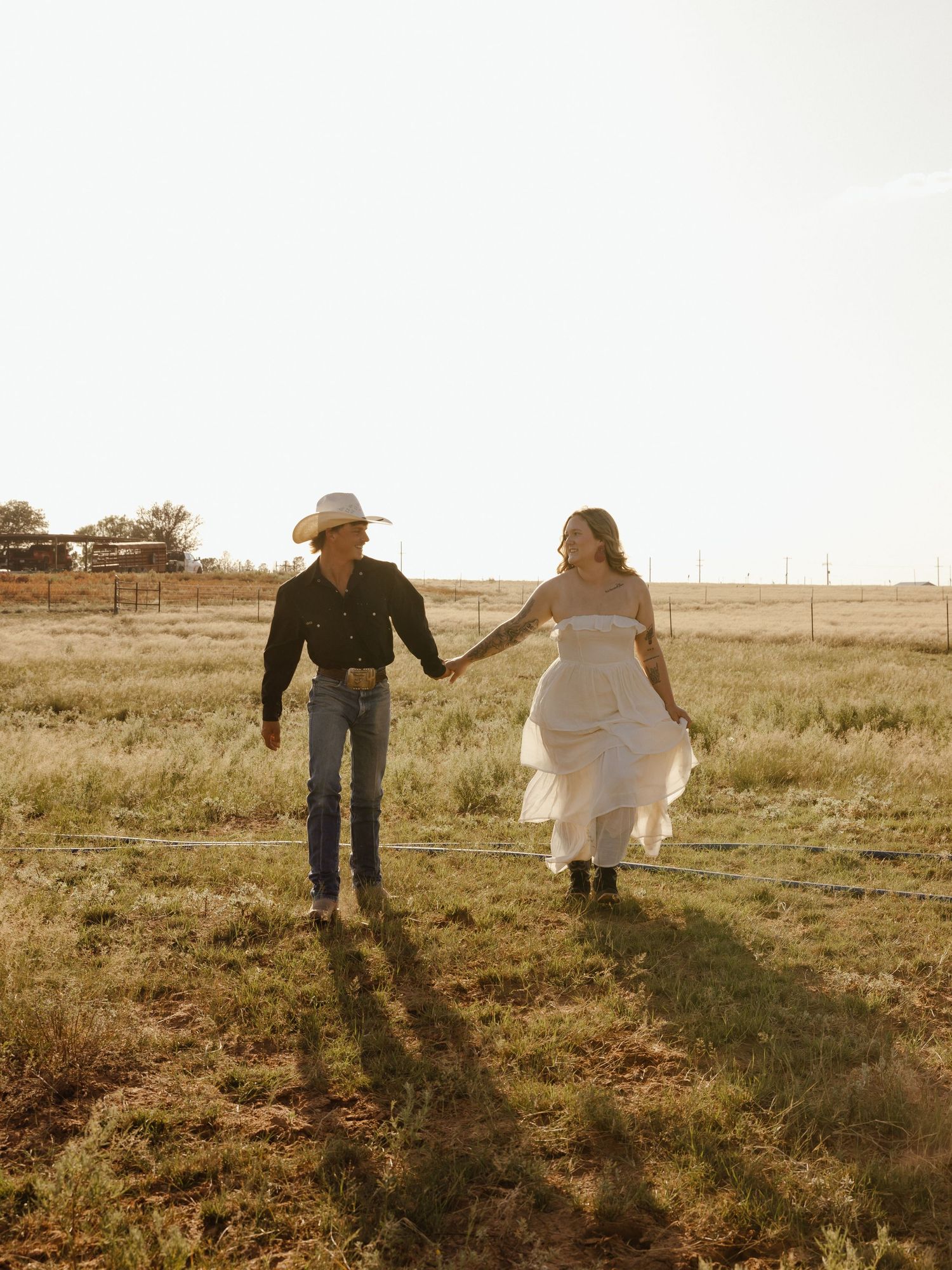 Couple in western attire holding hands walking through a golden sunlit ranch field at sunset.