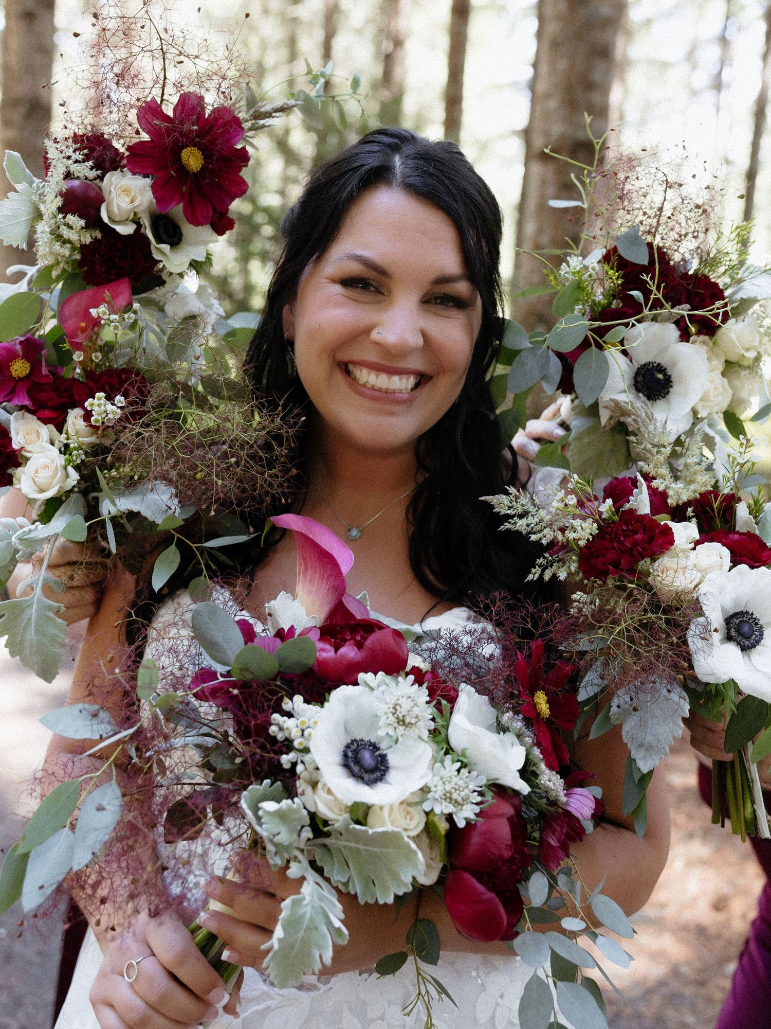 Smiling bride holding lush burgundy and white wedding bouquets with anemones and eucalyptus in a forest setting.