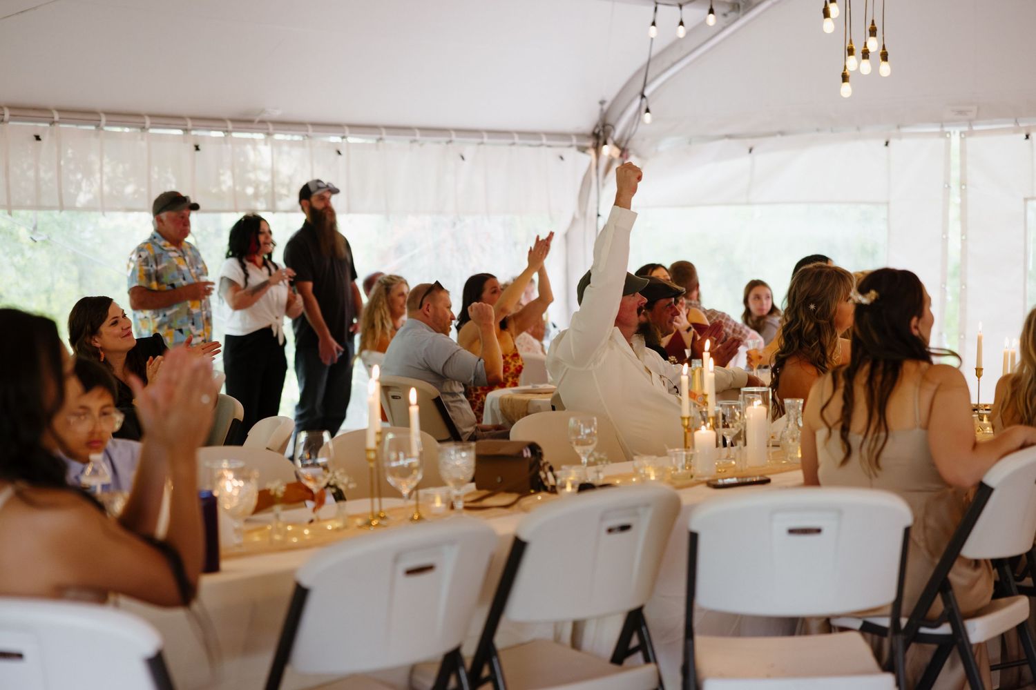 Lively wedding reception under white tent with guests celebrating, raising hands, seated at candlelit tables with string lights.