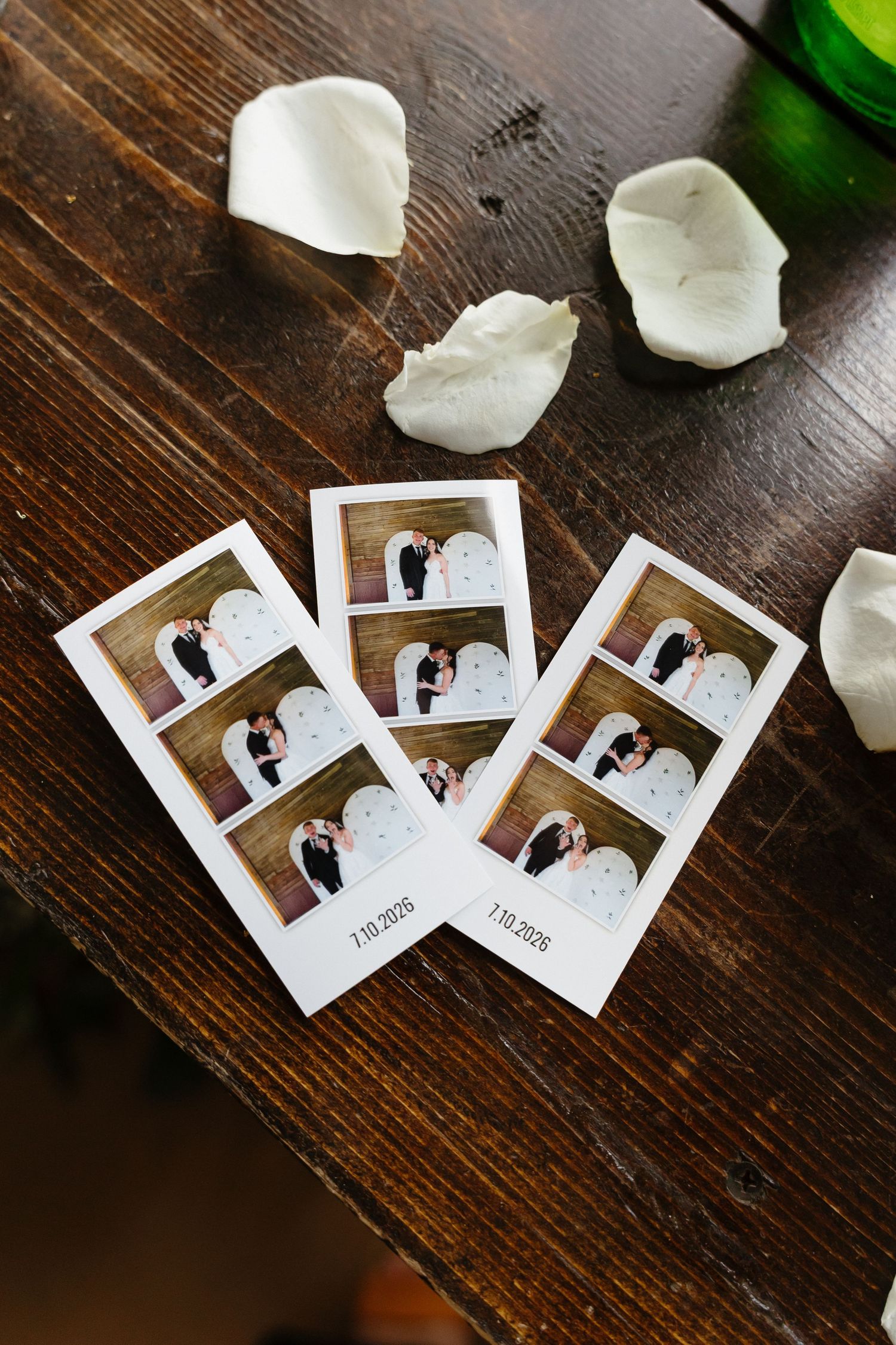 Three photo booth strips from wedding dated 7.19.2025 displayed on wooden table with rose petals.