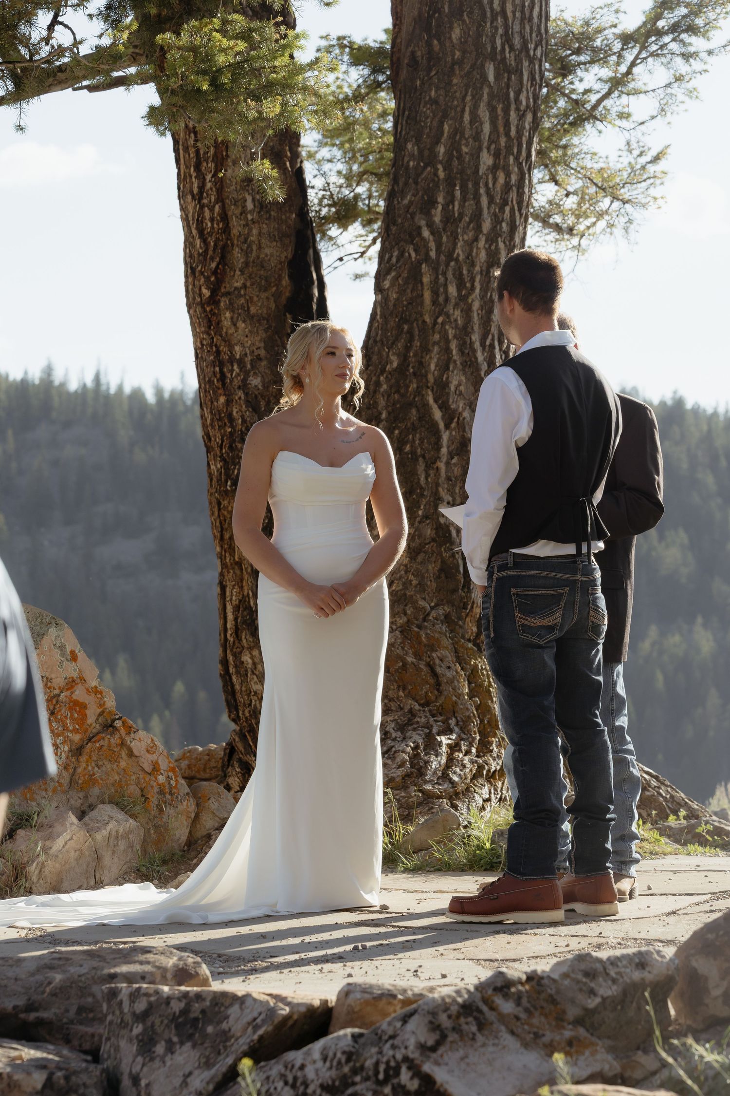 Bride in elegant white gown exchanging vows with groom outdoors among pine trees with mountain views.