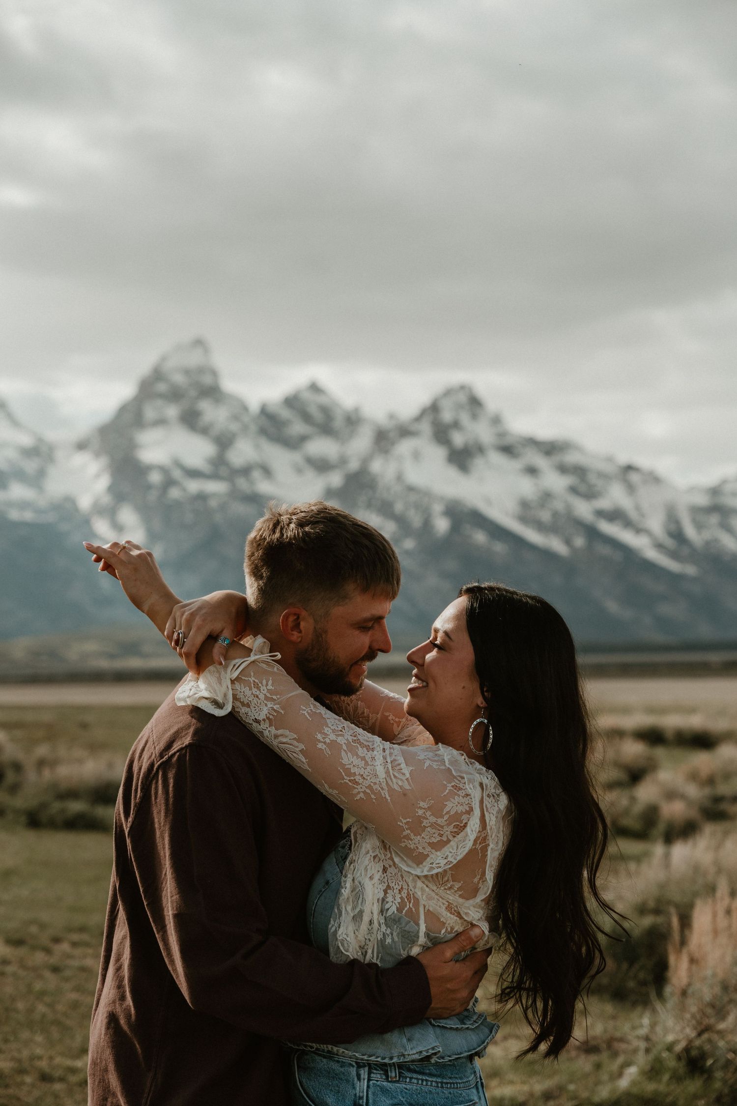 Smiling couple embracing and gazing at each other with the Grand Teton mountain range behind them.