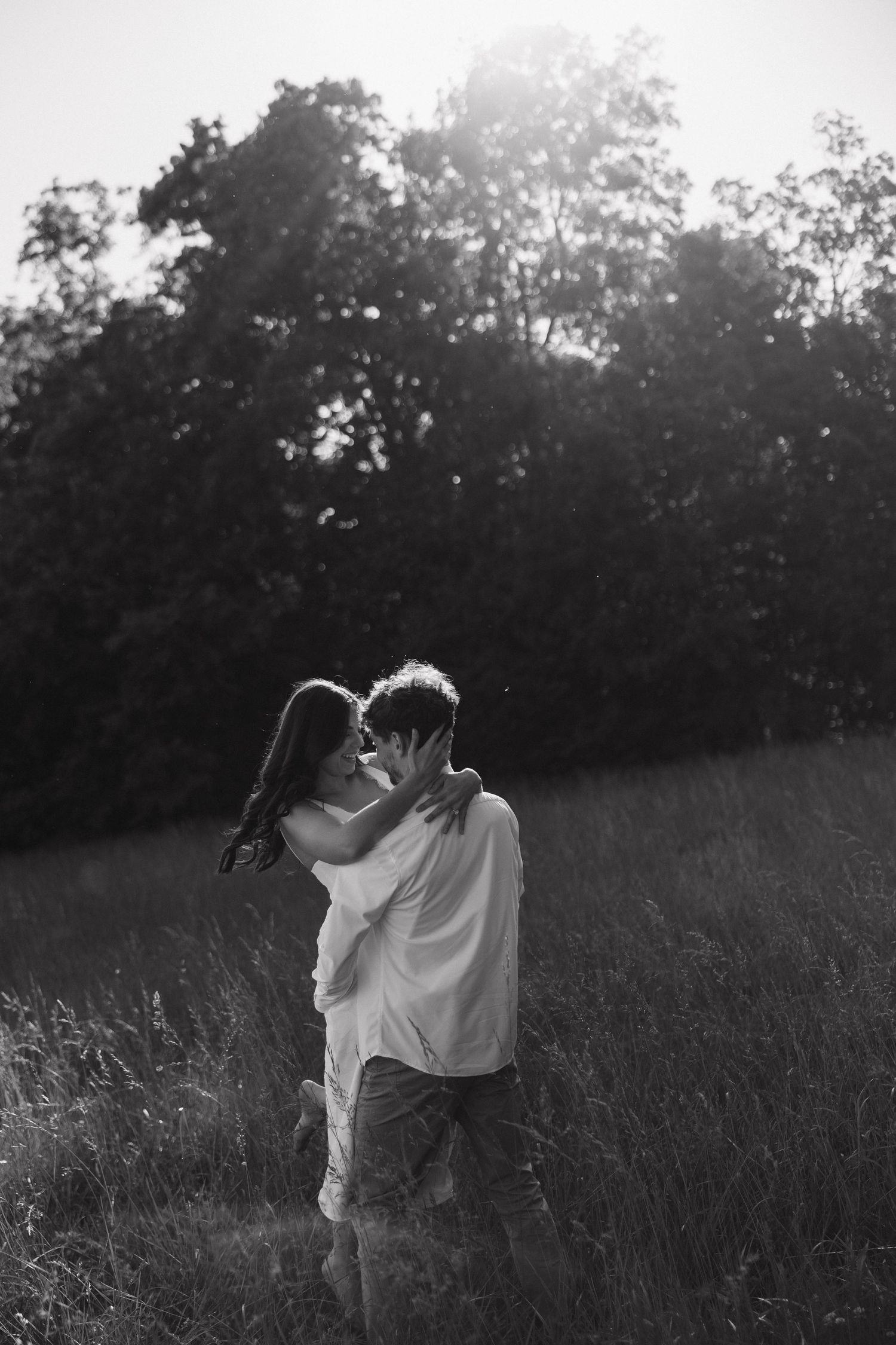 Black and white photo of a couple almost kissing in a sunny meadow with trees.