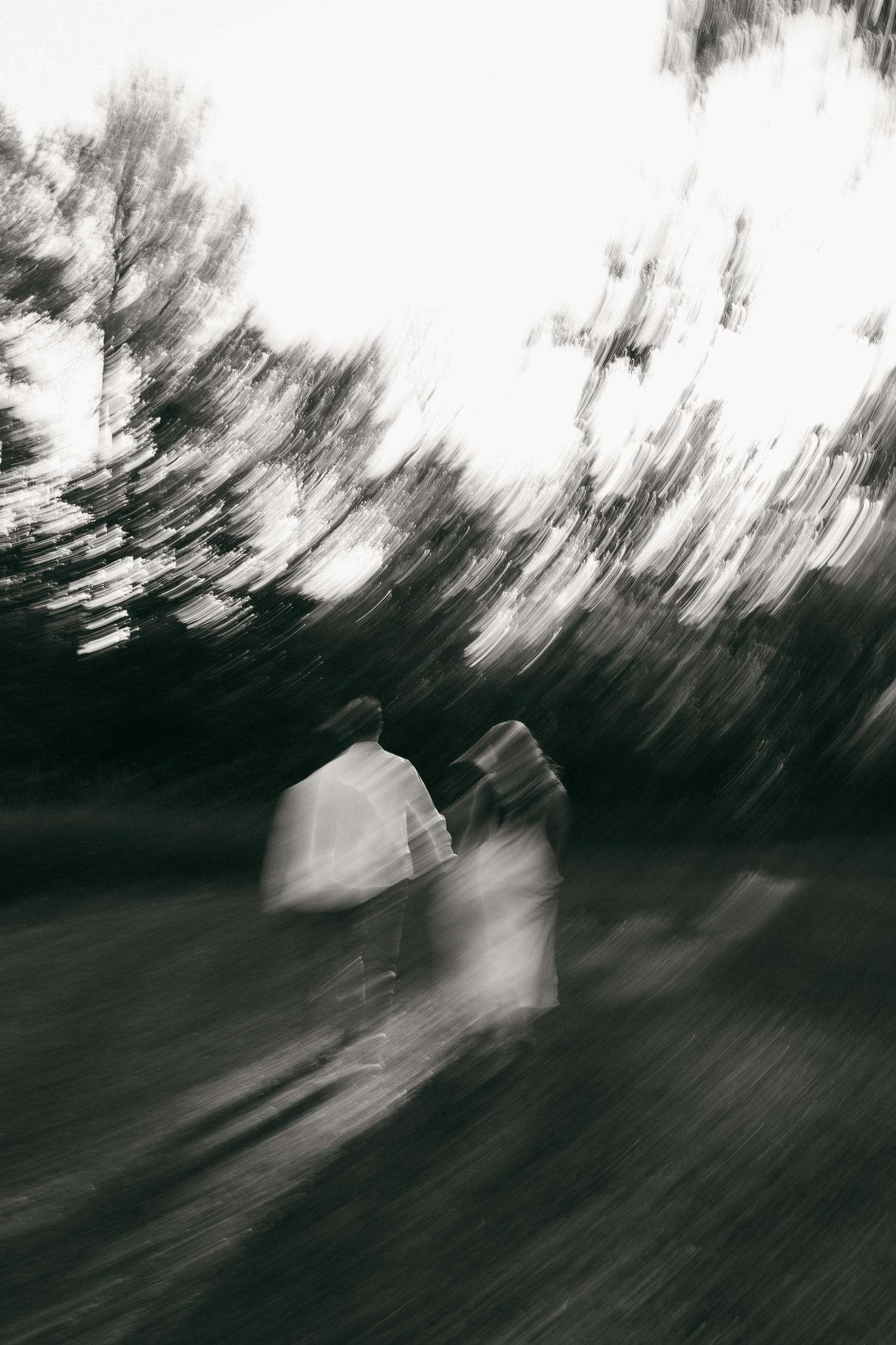 Black and white motion-blurred photo of a couple walking together through a tree-lined outdoor path.
