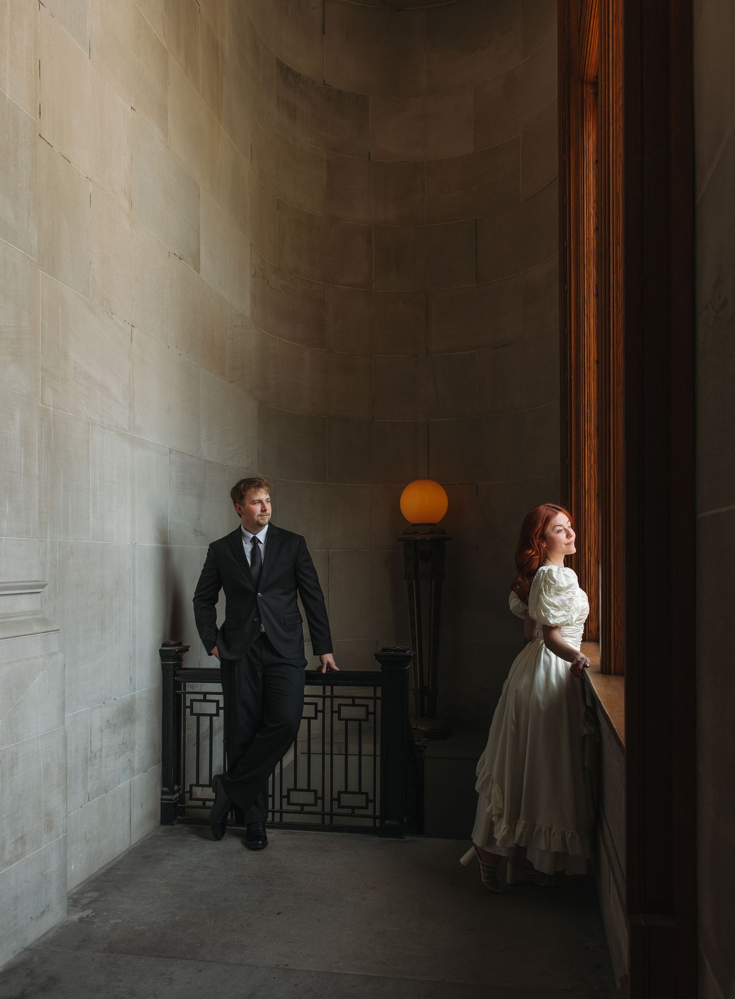 Bride in white dress and groom in black suit standing apart in a grand stone hallway with warm lighting.
