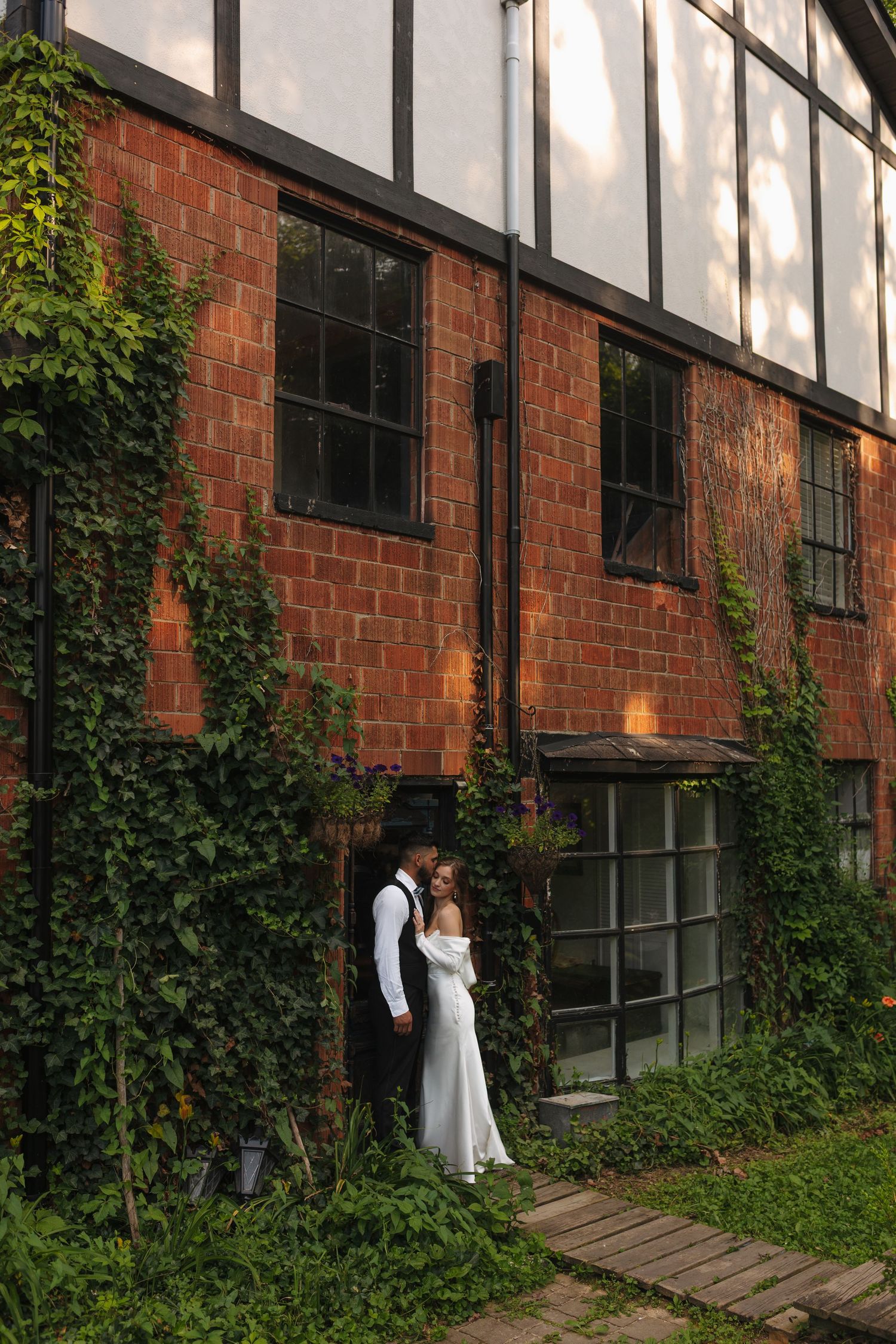 Bride and groom embracing by ivy-covered red brick building with large windows.