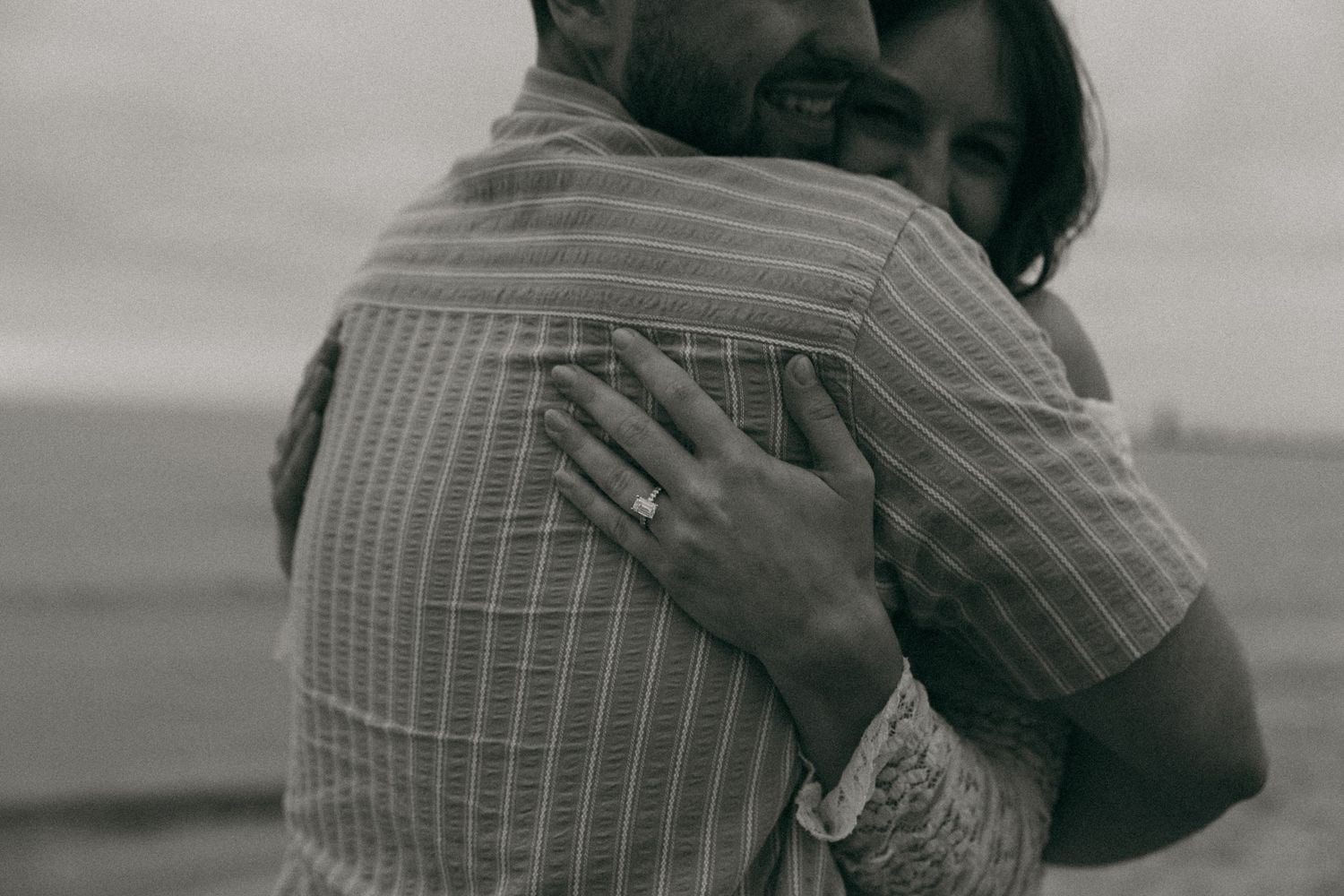 Black and white photo of woman hugging man showing engagement ring on her hand.