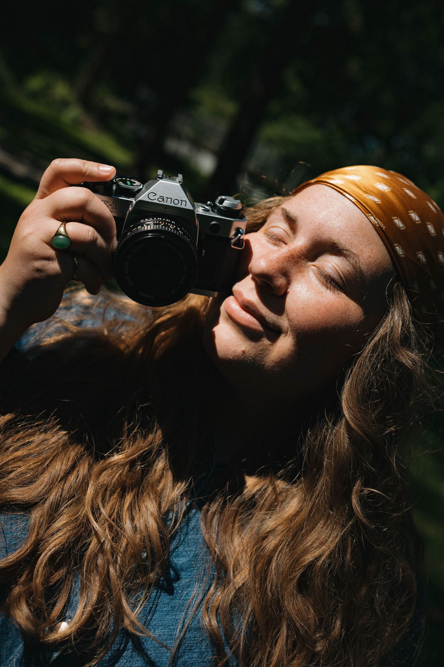 Young woman with curly hair and yellow bandana holding Canon film camera in sunlight.