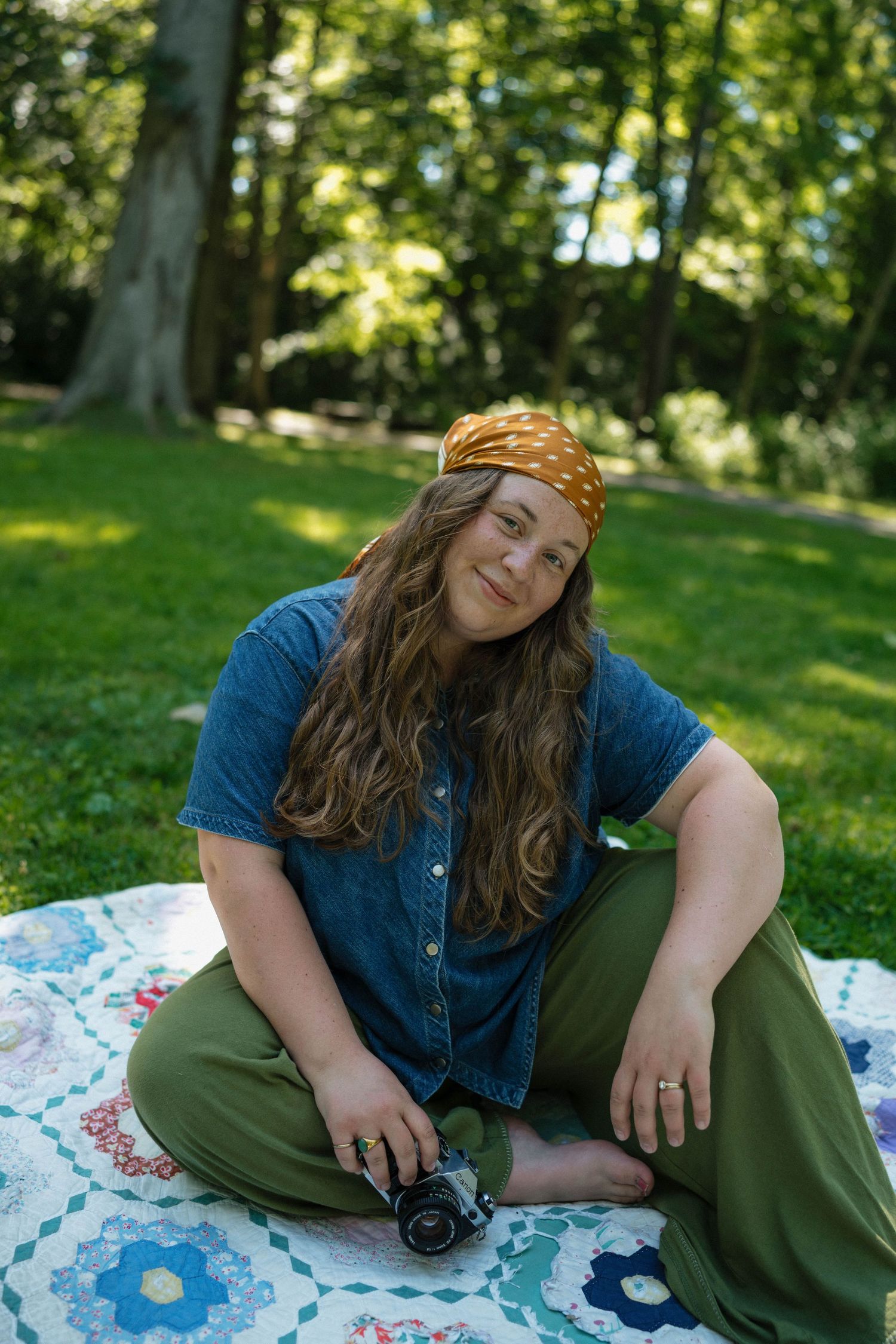 Young woman with curly hair and orange bandana sitting on quilt in park holding vintage camera.