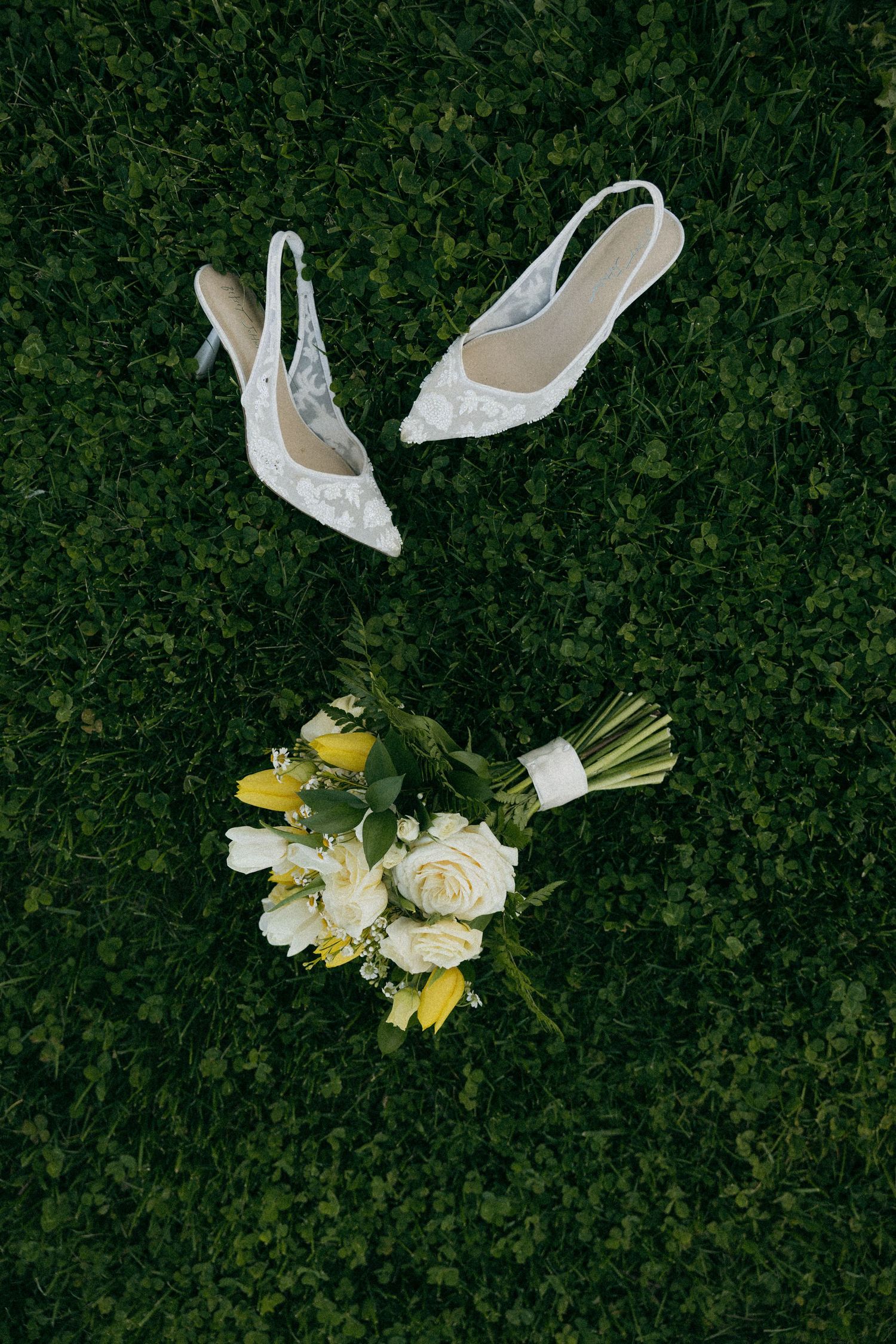 White lace bridal heels and a yellow and white wedding bouquet arranged on lush green grass.