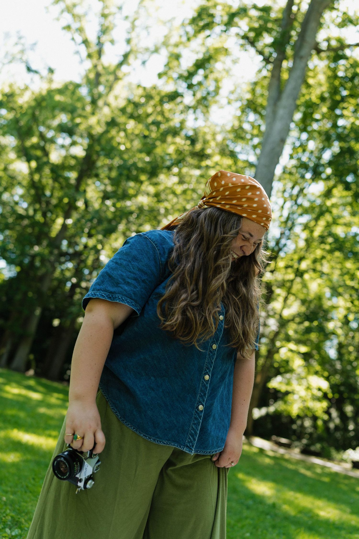 Young woman in denim shirt and mustard bandana holding film camera while looking down in park.