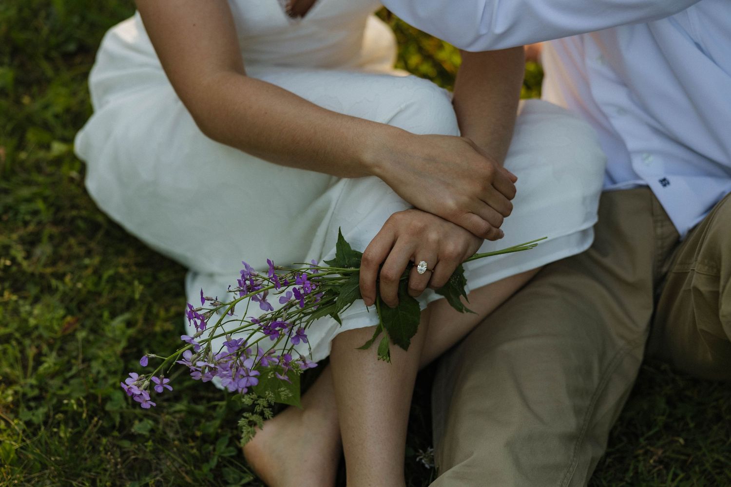 Couple sitting on grass holding purple wildflowers with engagement ring visible.