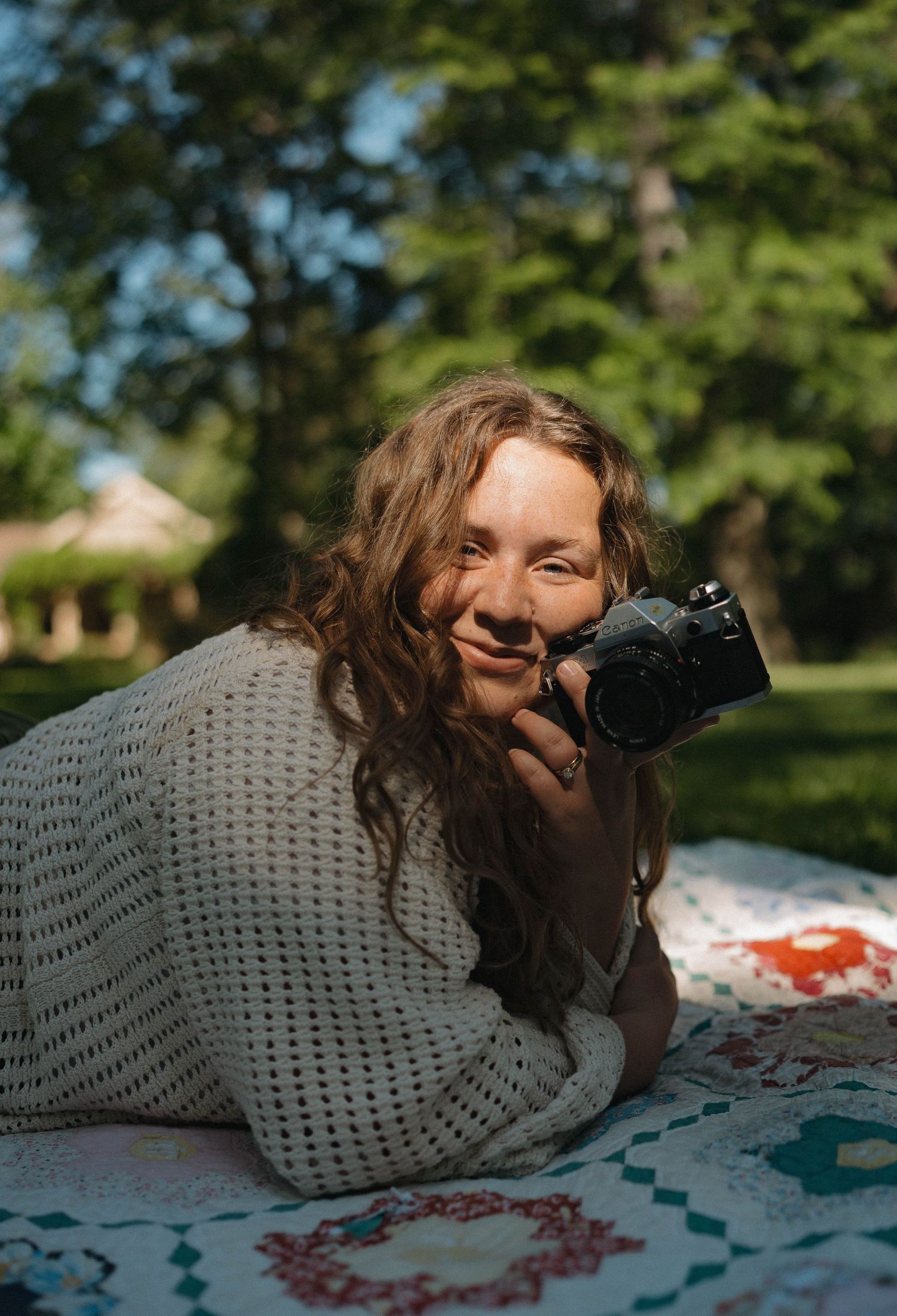Smiling woman lying on quilt in park holding vintage film camera close to her face.