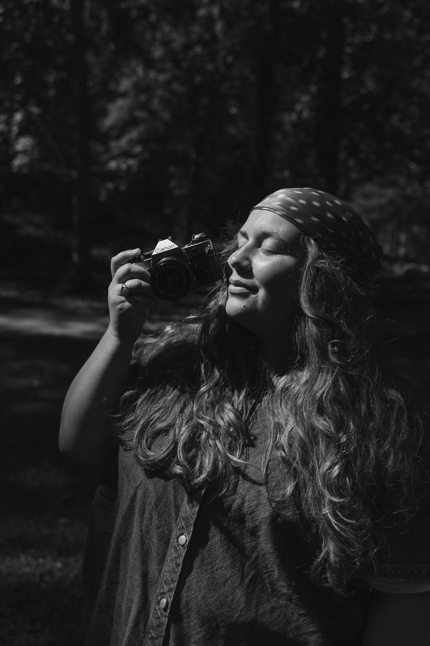 Black and white portrait of a smiling woman with long hair holding a film camera outdoors.