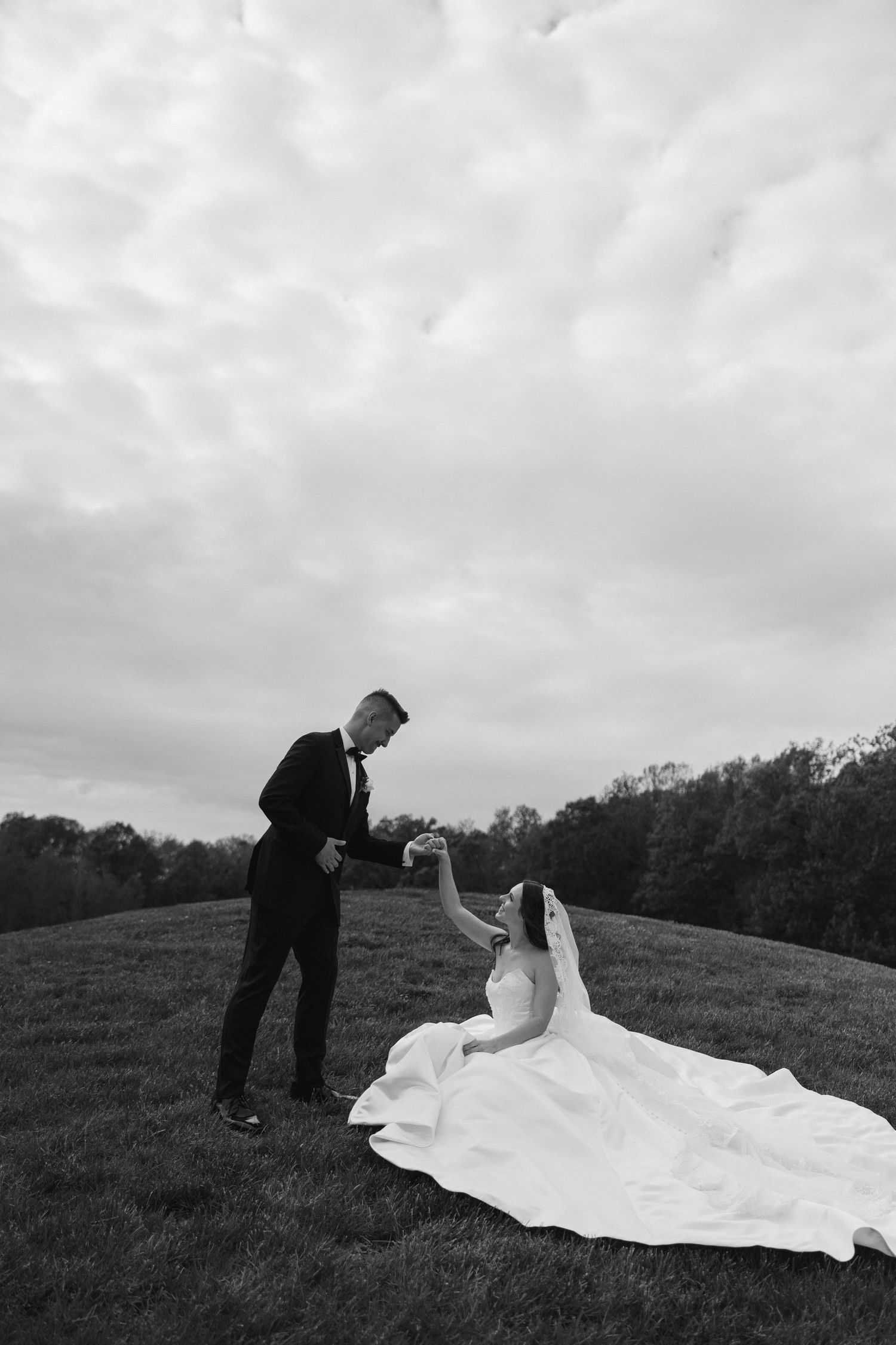 Black and white photo of a bride sitting on a hill as the groom holds her hand outdoors.