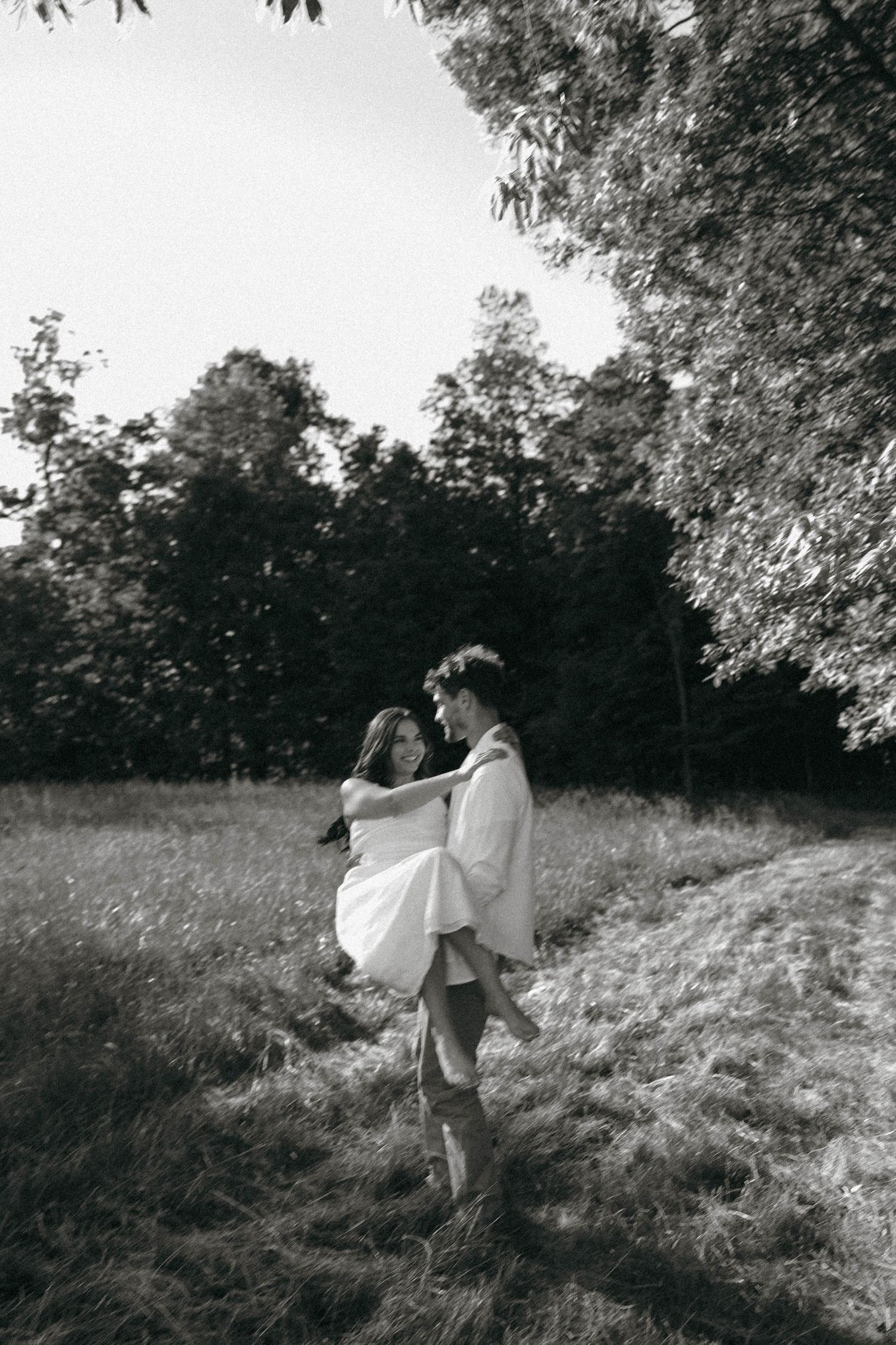 Black and white photo of groom lifting bride in a field surrounded by trees.