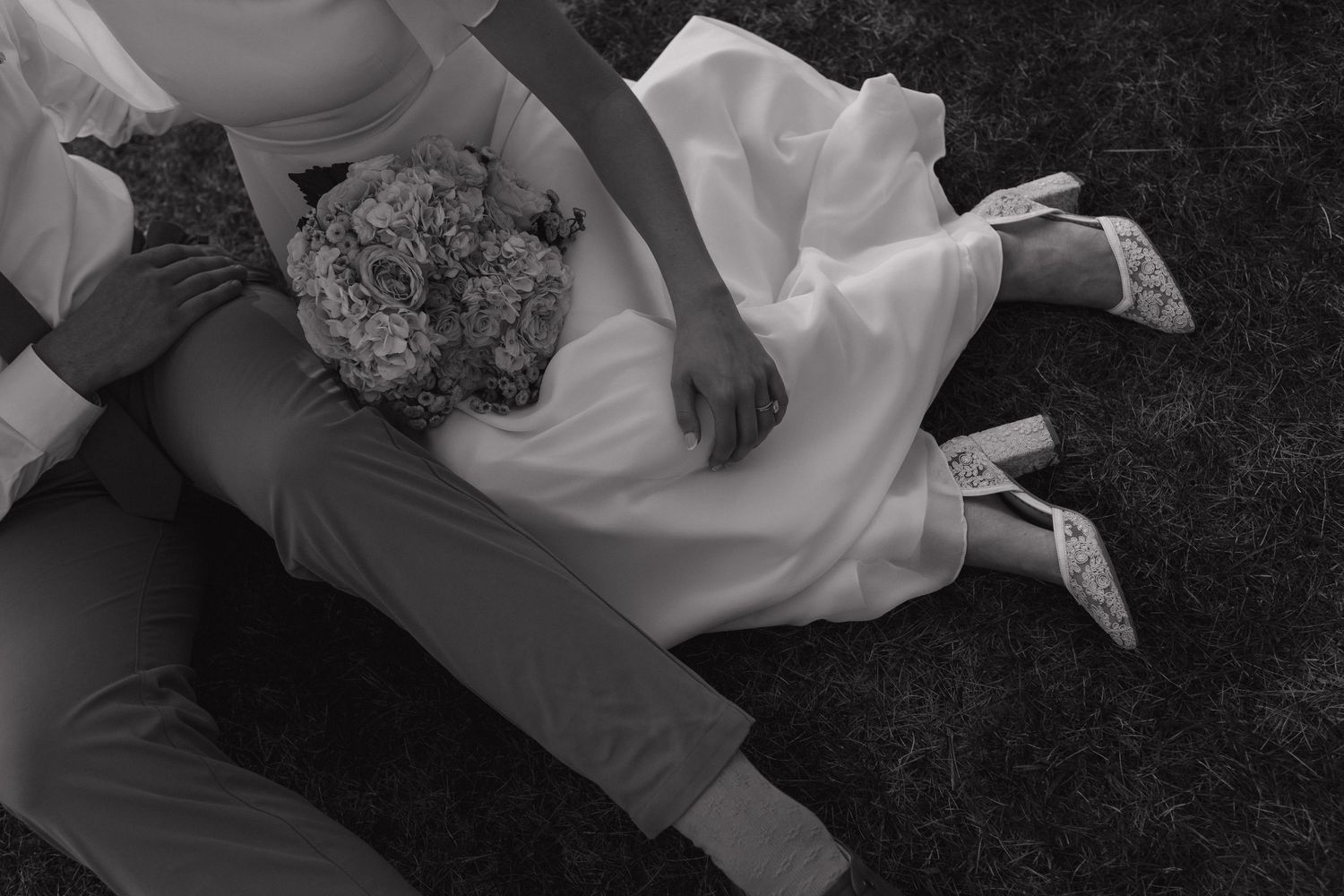 Black and white photo of bride and groom sitting on grass with wedding bouquet.