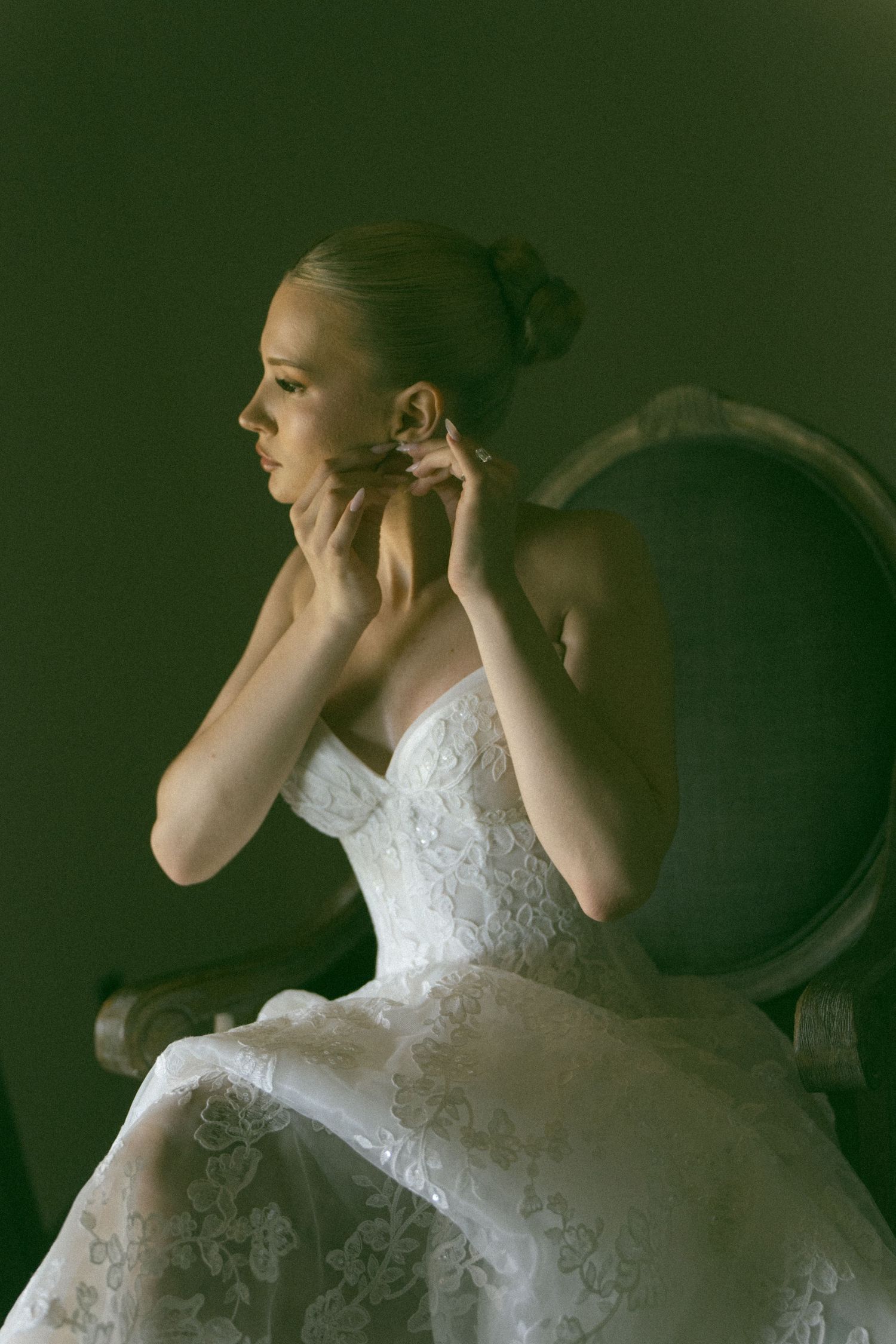 Bride in white lace strapless gown sitting in vintage chair, putting on earrings against dark background.
