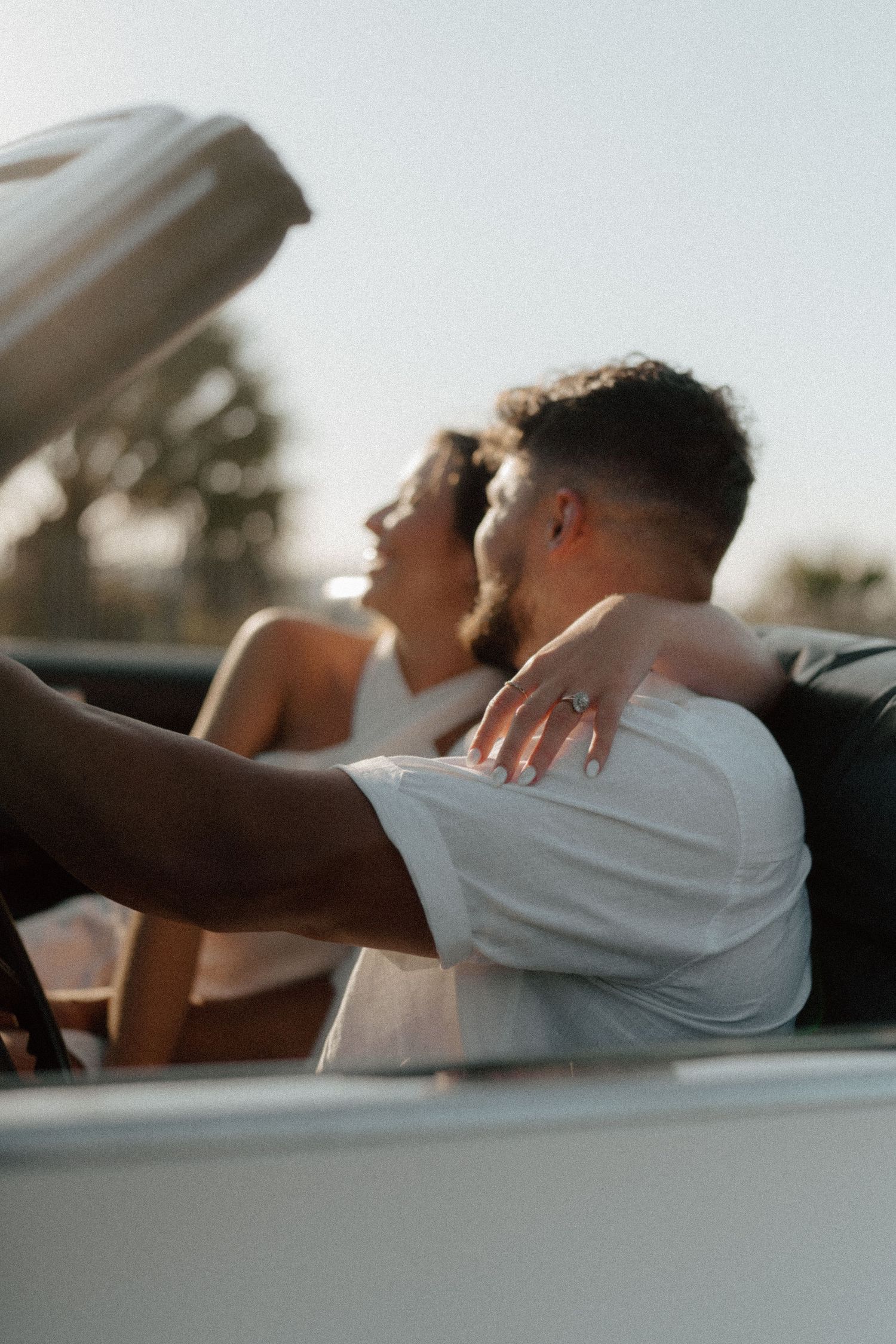 Couple sitting in a convertible car, woman placing her hand on man's shoulder, enjoying a sunny day outdoors.