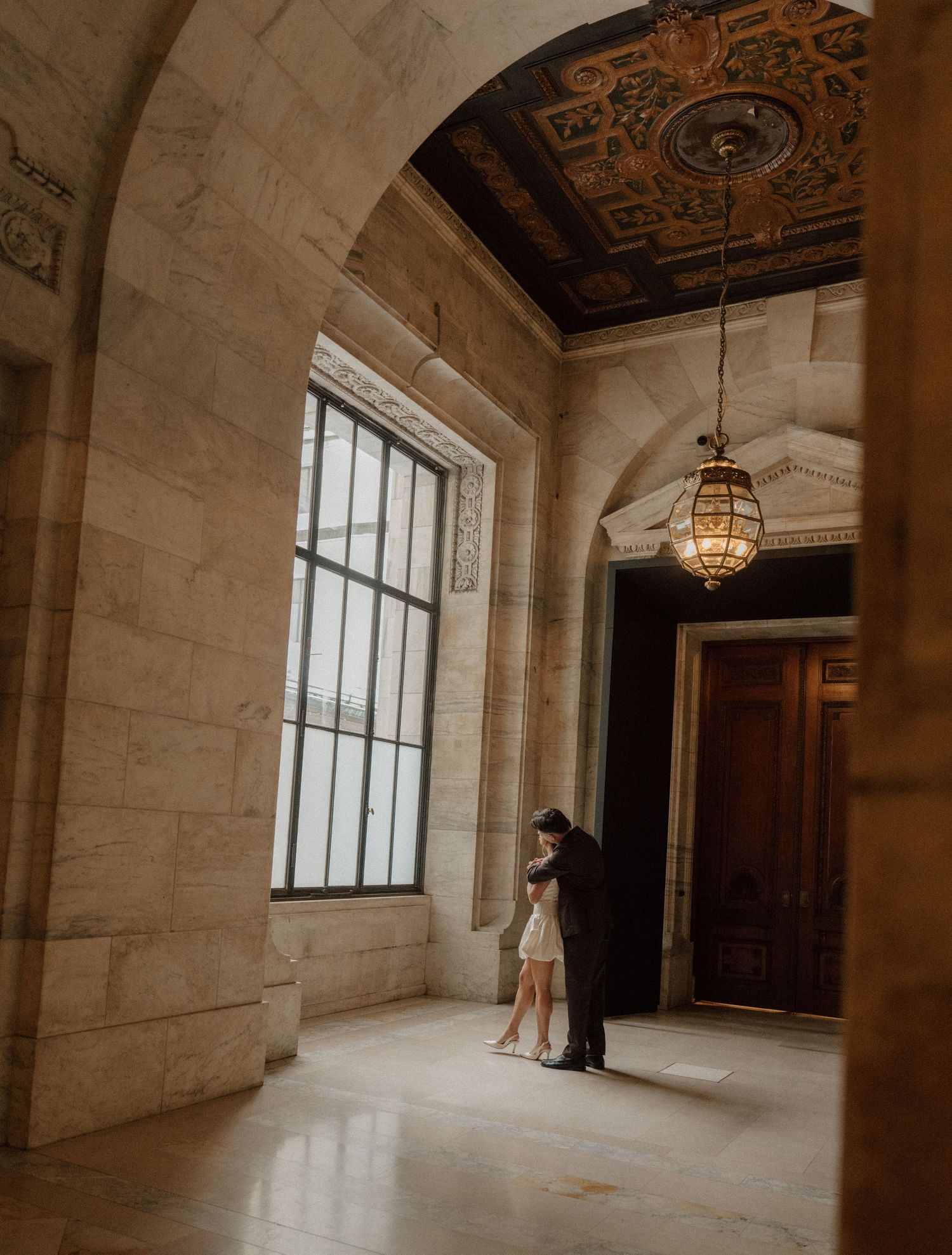 Couple embracing in elegant marble hallway with ornate ceiling and vintage lantern in historic building.