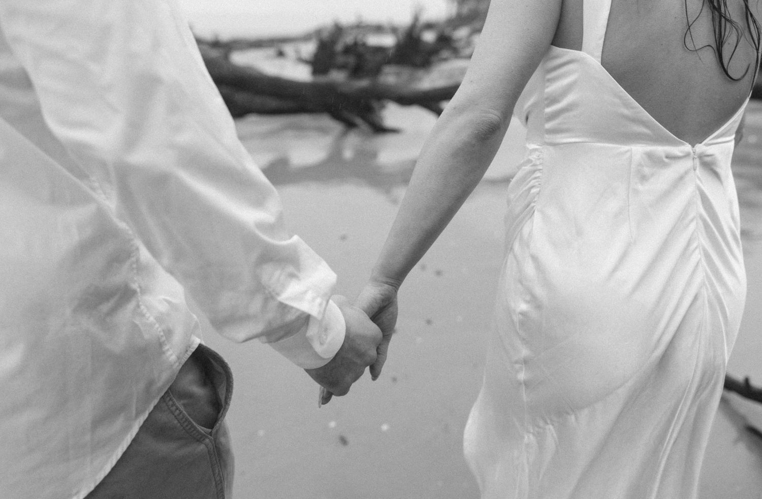Black and white photo of a couple holding hands on a beach, woman in elegant backless white wedding gown. Jekyll Island Engagement