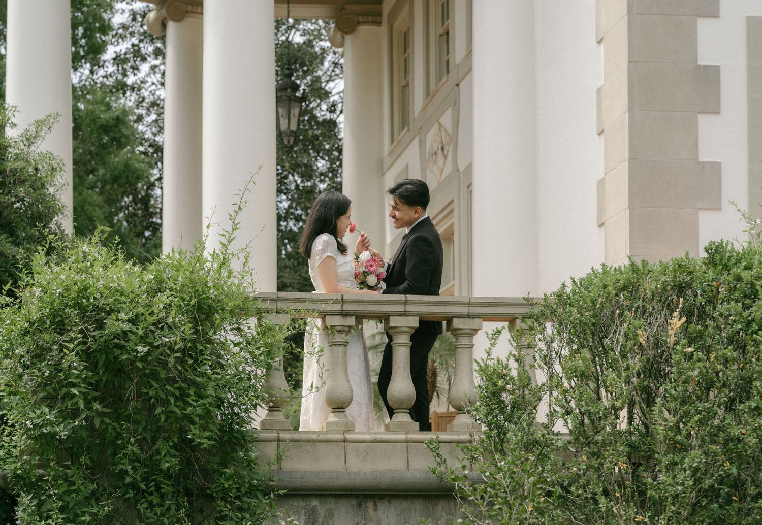 Couple in wedding attire exchanging vows on elegant balcony with white columns and lush greenery.