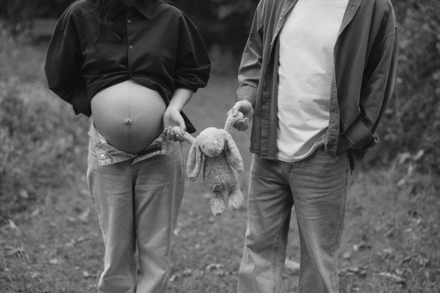 Black and white photo of pregnant couple holding stuffed bunny outdoors, maternity portrait in nature.