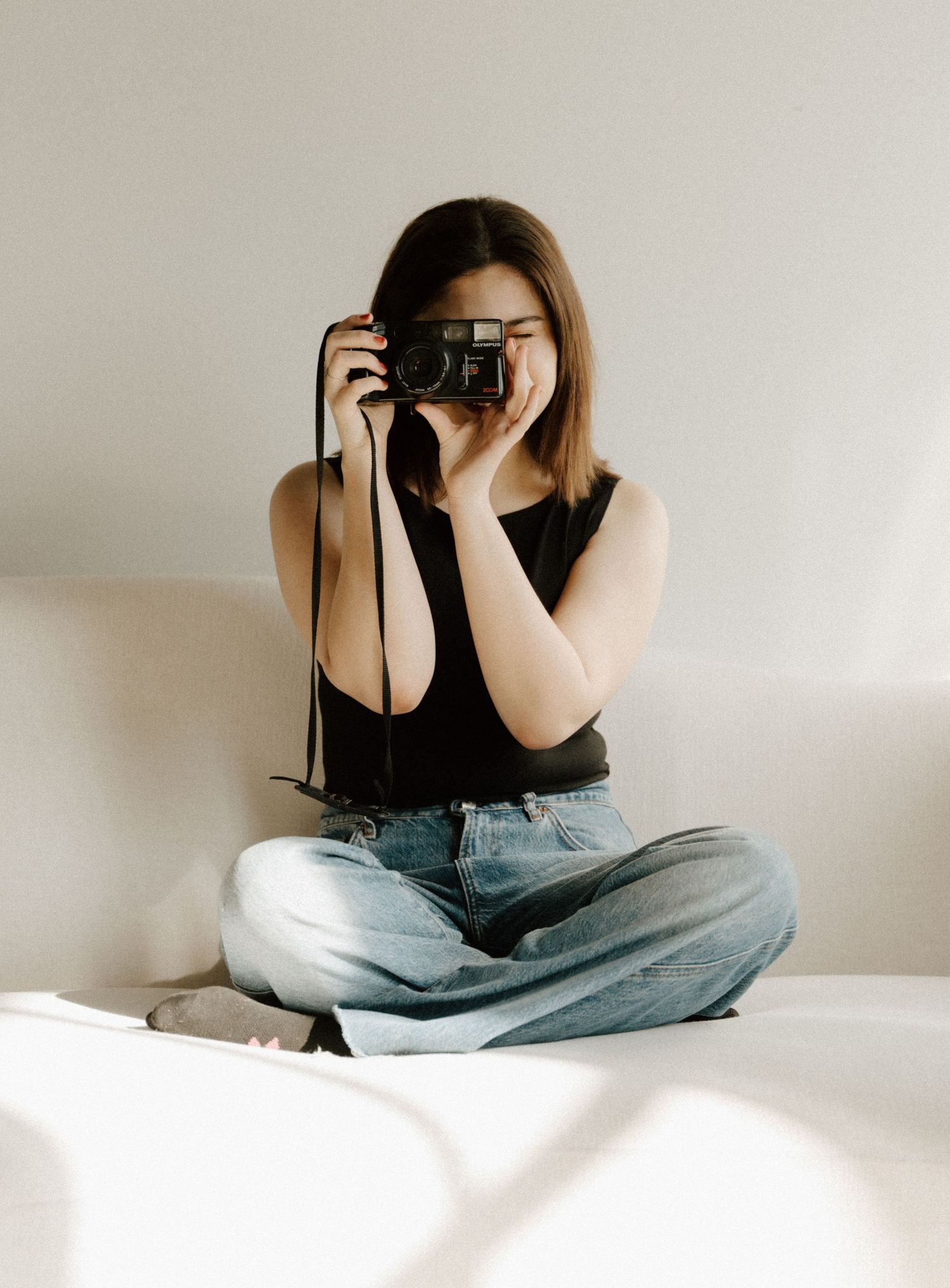 Young woman sitting cross-legged on a couch, holding a black film camera up to her face, wearing jeans and a black tank top.