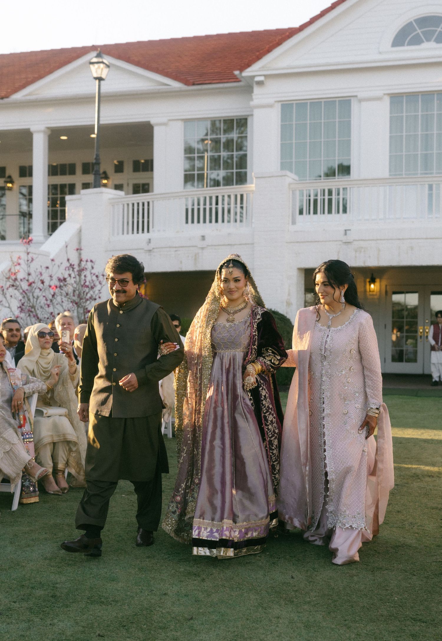 A bride in traditional South Asian attire walks with two escorts toward a white colonial venue.