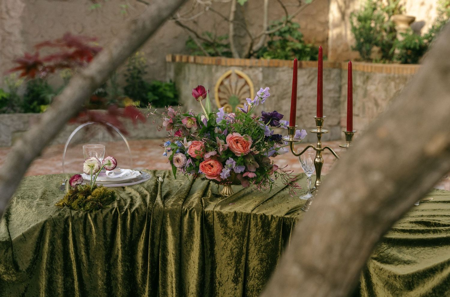 Elegant outdoor wedding tablescape with green velvet linen, pink floral centerpiece, and red taper candles in a garden setting.
