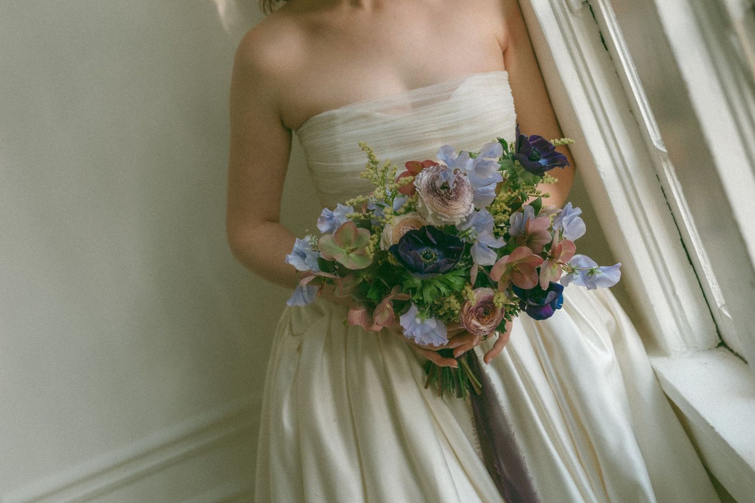 Bride in strapless ivory gown holding lush blue and pink floral bouquet with anemones near a sunlit window.
