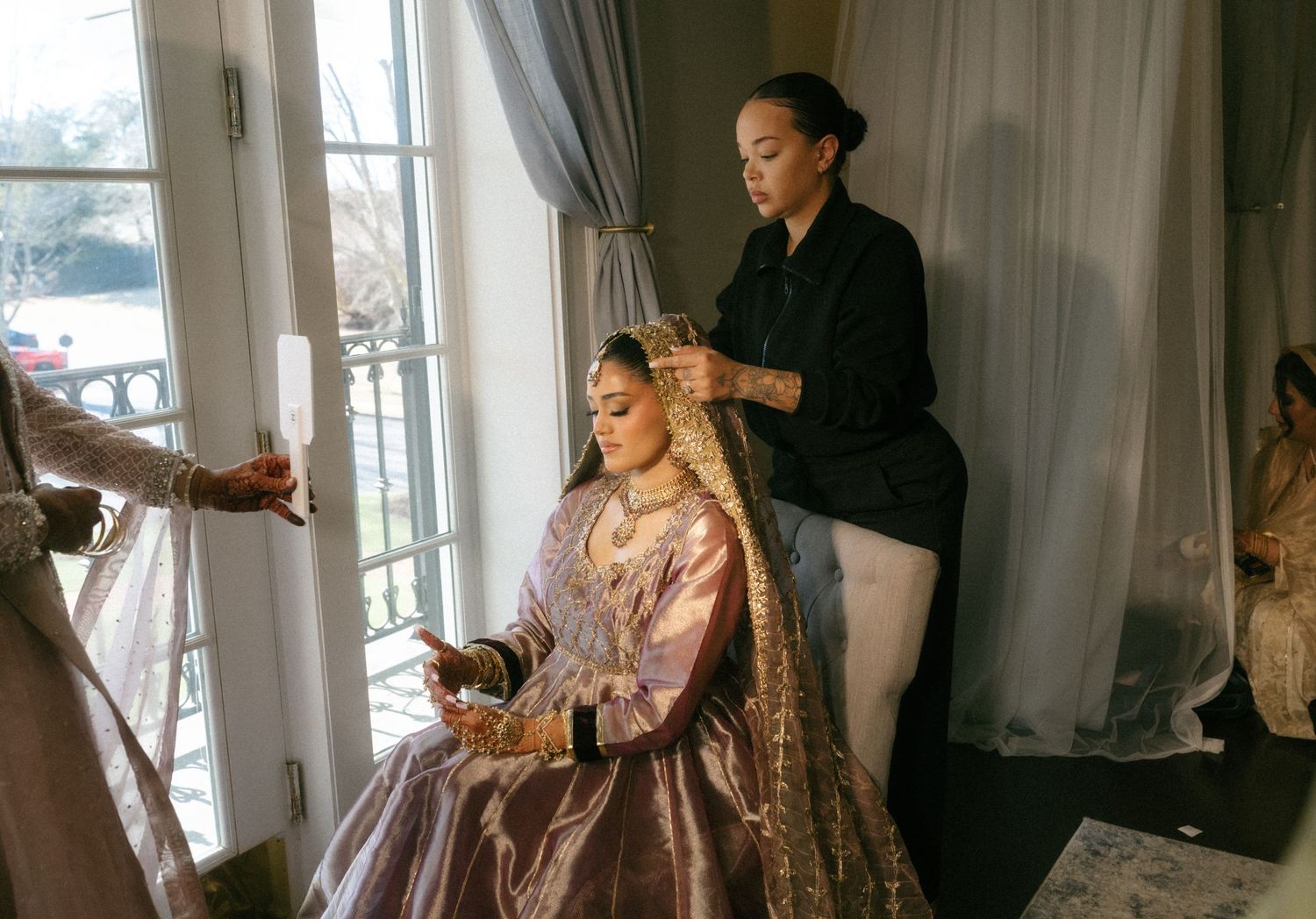 A bride in an ornate pink lehenga sits as a stylist arranges her hair near a bright window during bridal preparations.