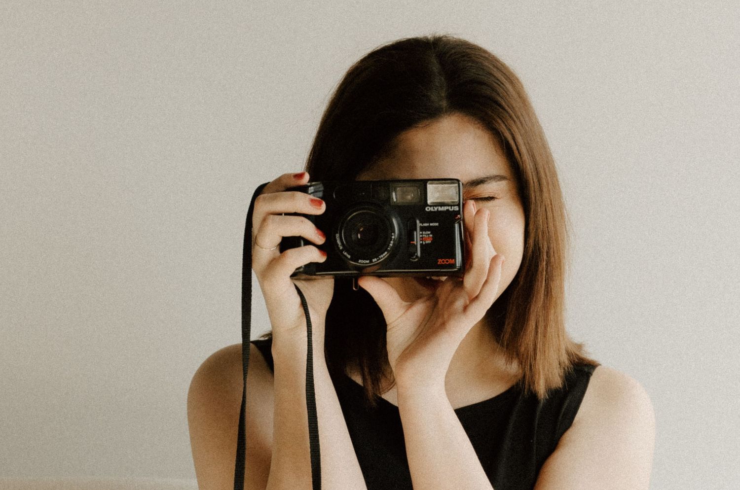 A young woman holds a vintage black film camera up to her face, wearing a sleeveless black top against a neutral background.