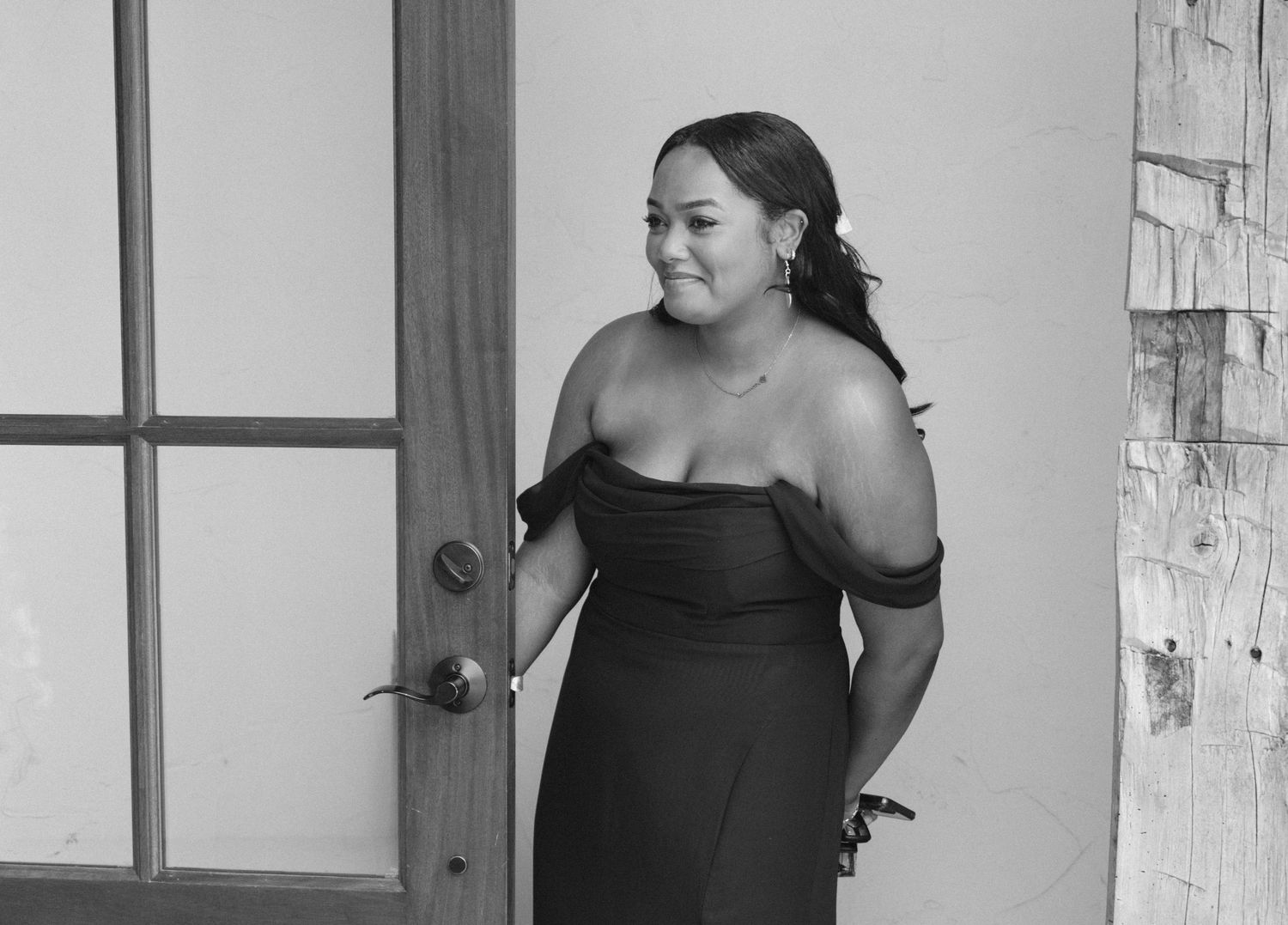 Woman in black off-shoulder gown smiling near glass door in elegant black and white portrait photo.