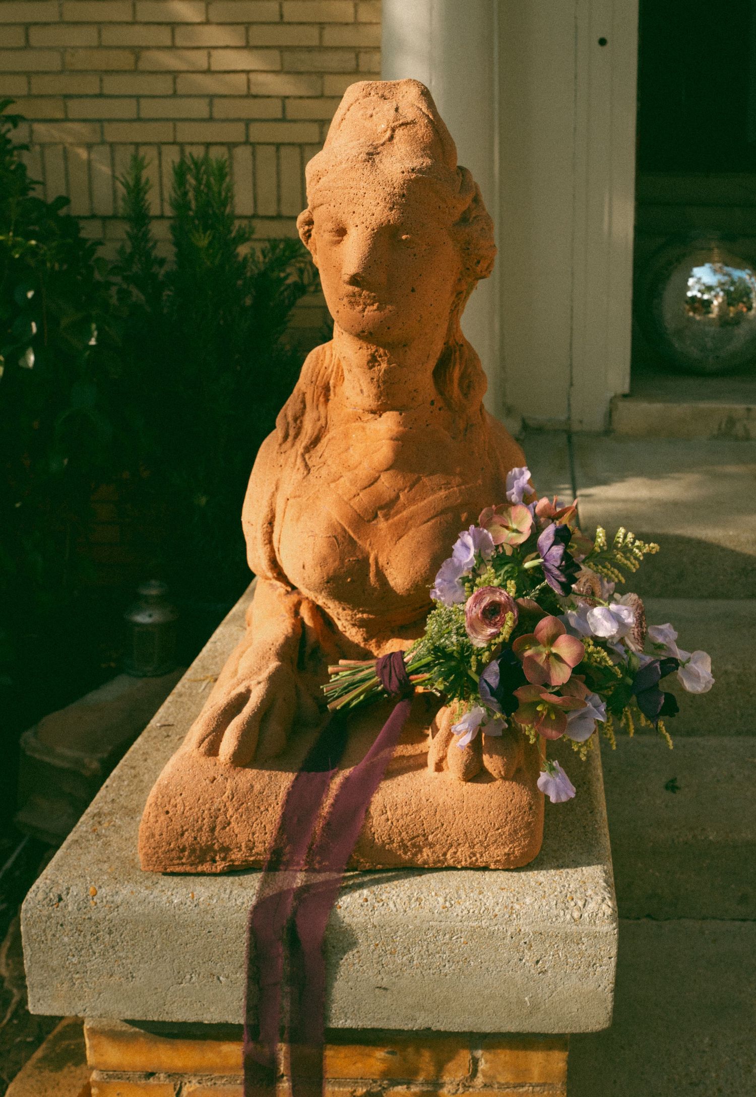 Rustic terracotta sphinx sculpture with purple and pink bridal bouquet tied with ribbon on stone pedestal.