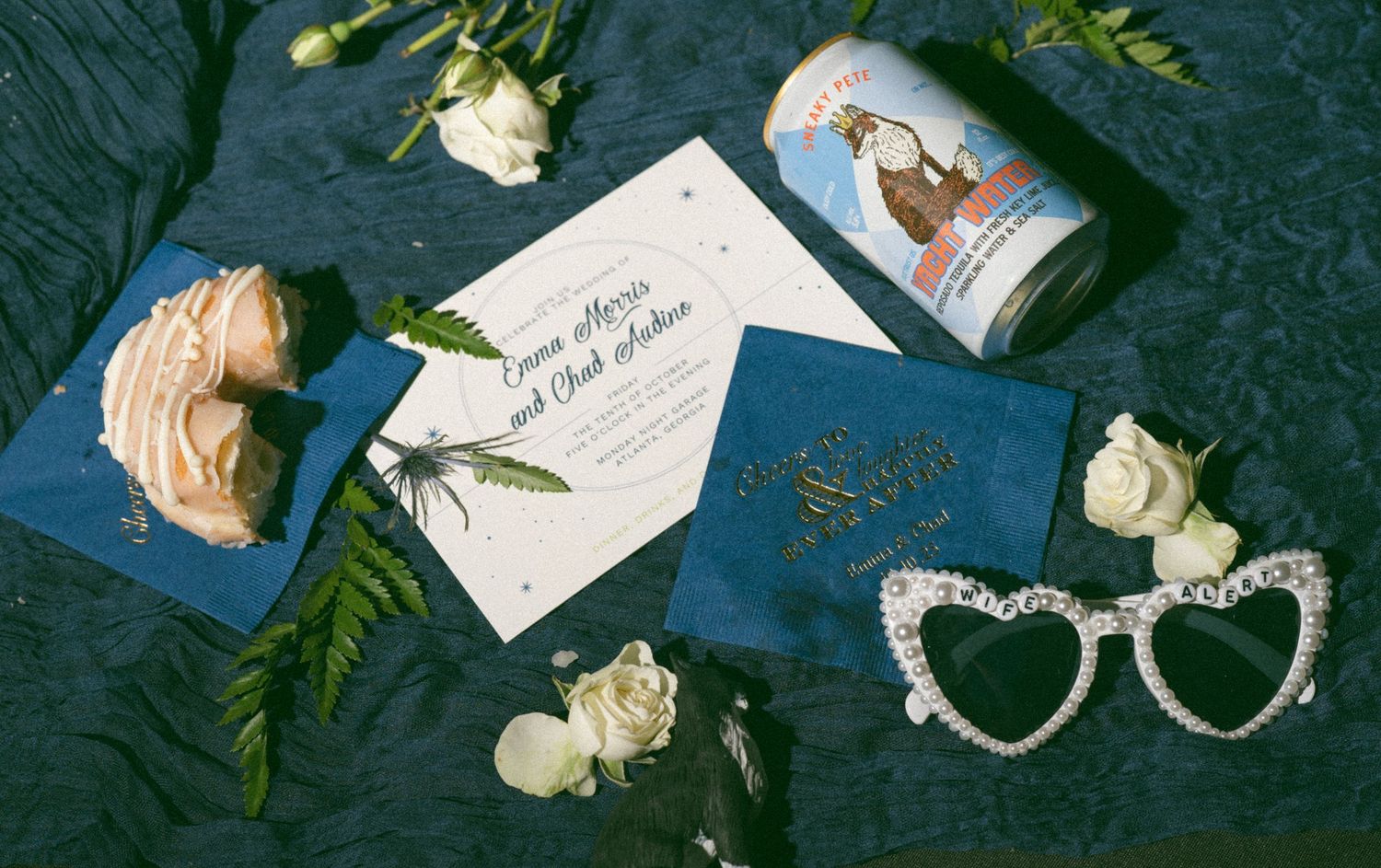 Wedding invitation suite with white roses, pearl heart sunglasses, navy envelopes, and a vintage beer can on dark background.