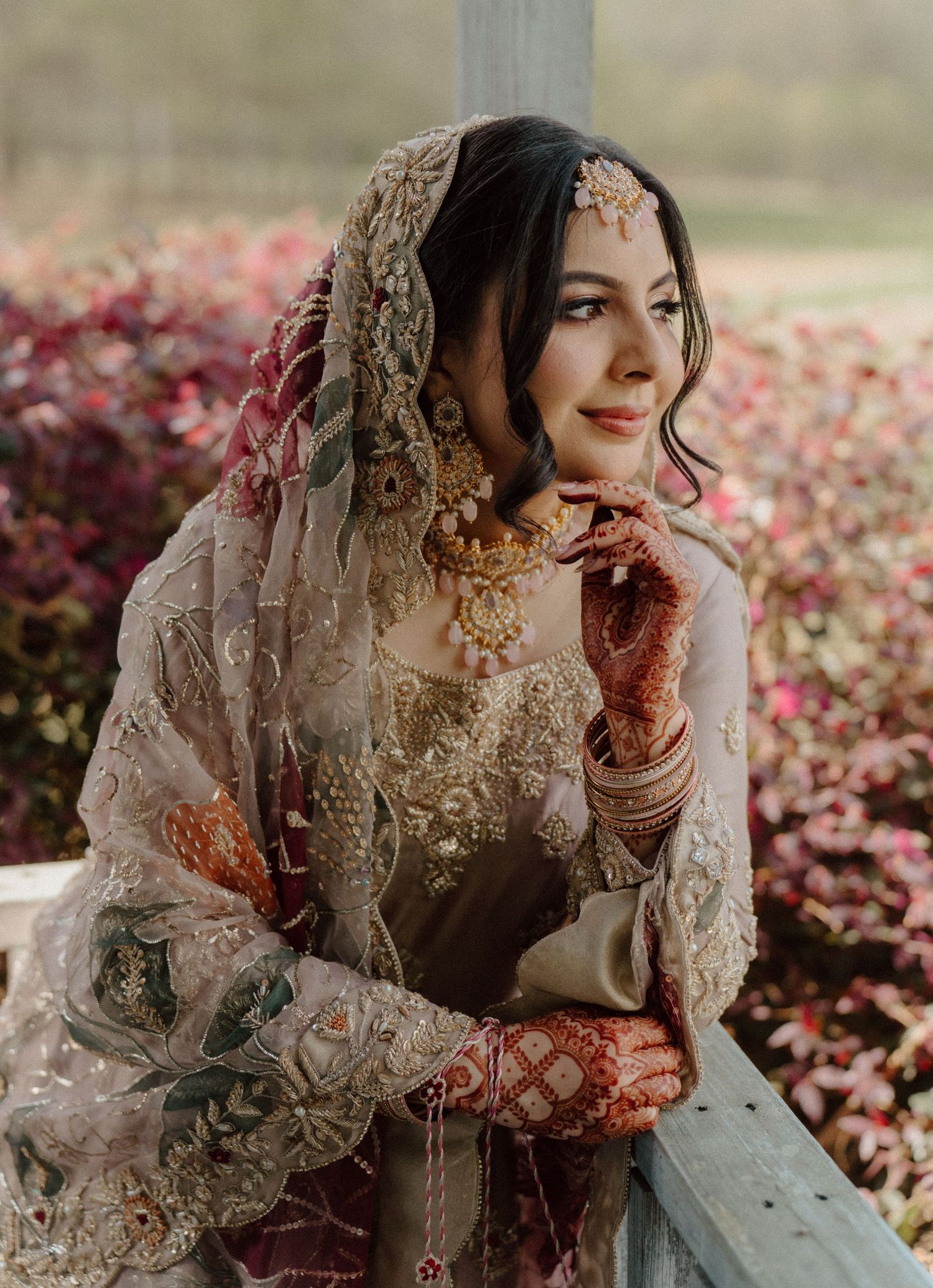 A bride in traditional South Asian bridal attire with gold jewelry, mehndi, and embroidered dupatta poses gracefully outdoors.