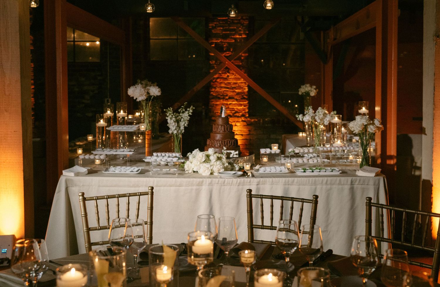 Elegant wedding reception table with white floral arrangements, candles, and a tiered cake in a warmly lit venue.