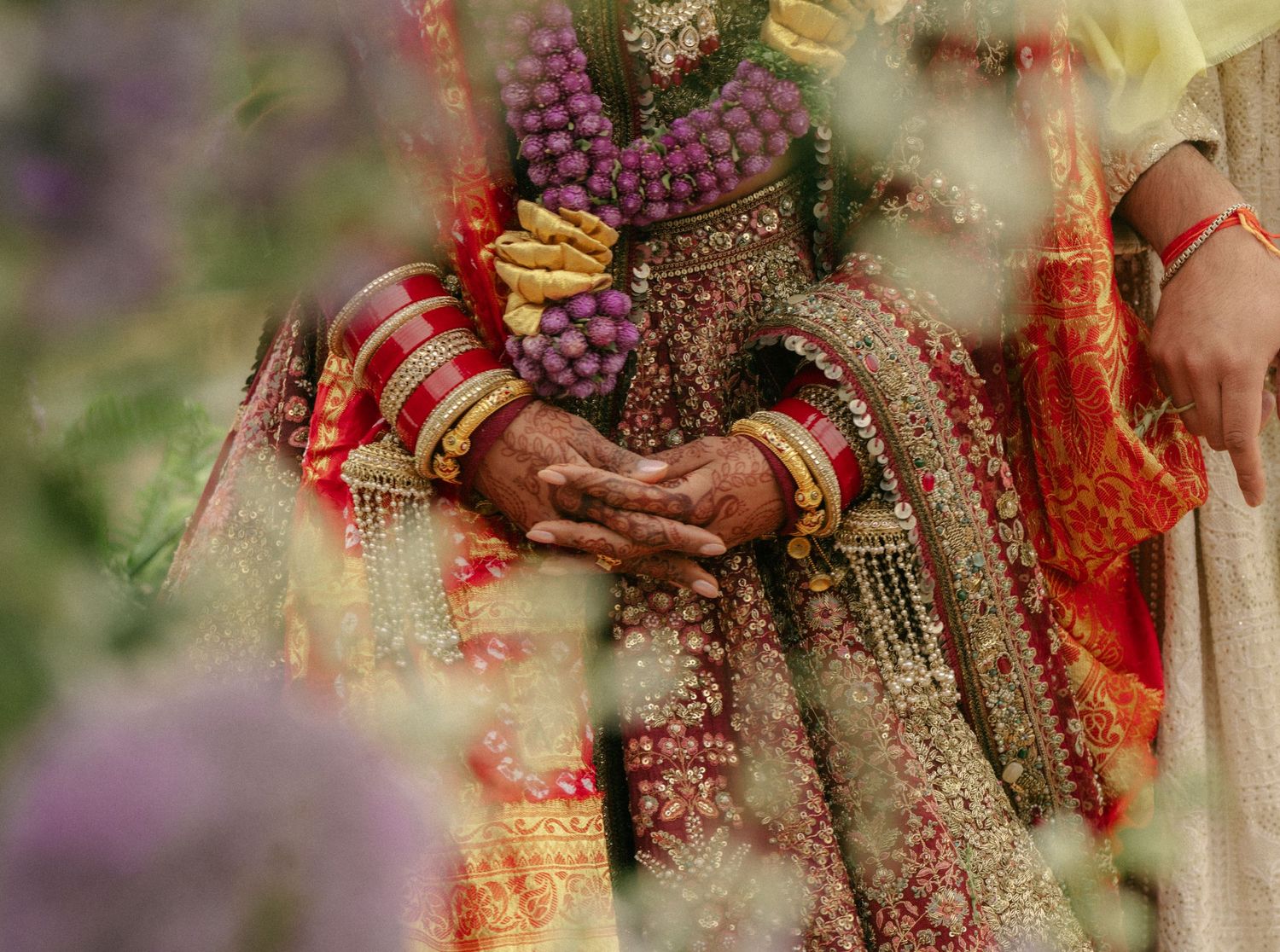 Close-up of Indian bride and groom hands adorned with bangles, mehndi, and floral garlands during traditional wedding ceremony.