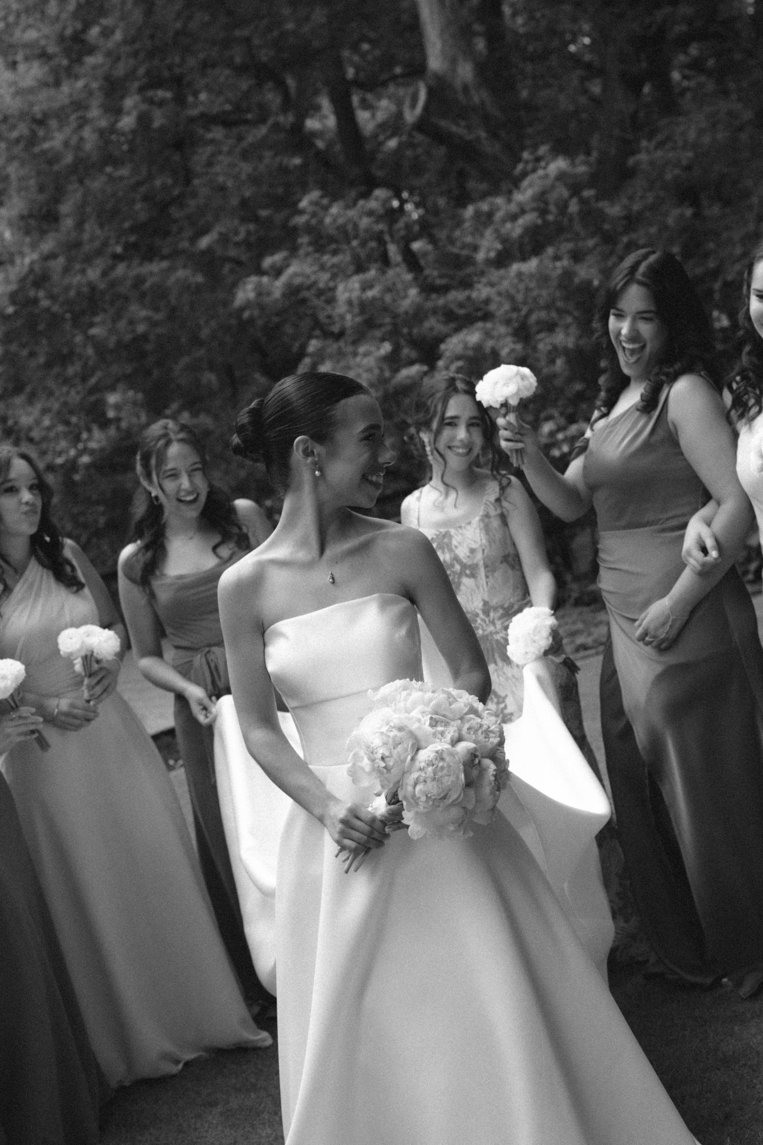Bride laughing with bridesmaids outdoors, holding bouquet in white gown, black and white wedding photo.