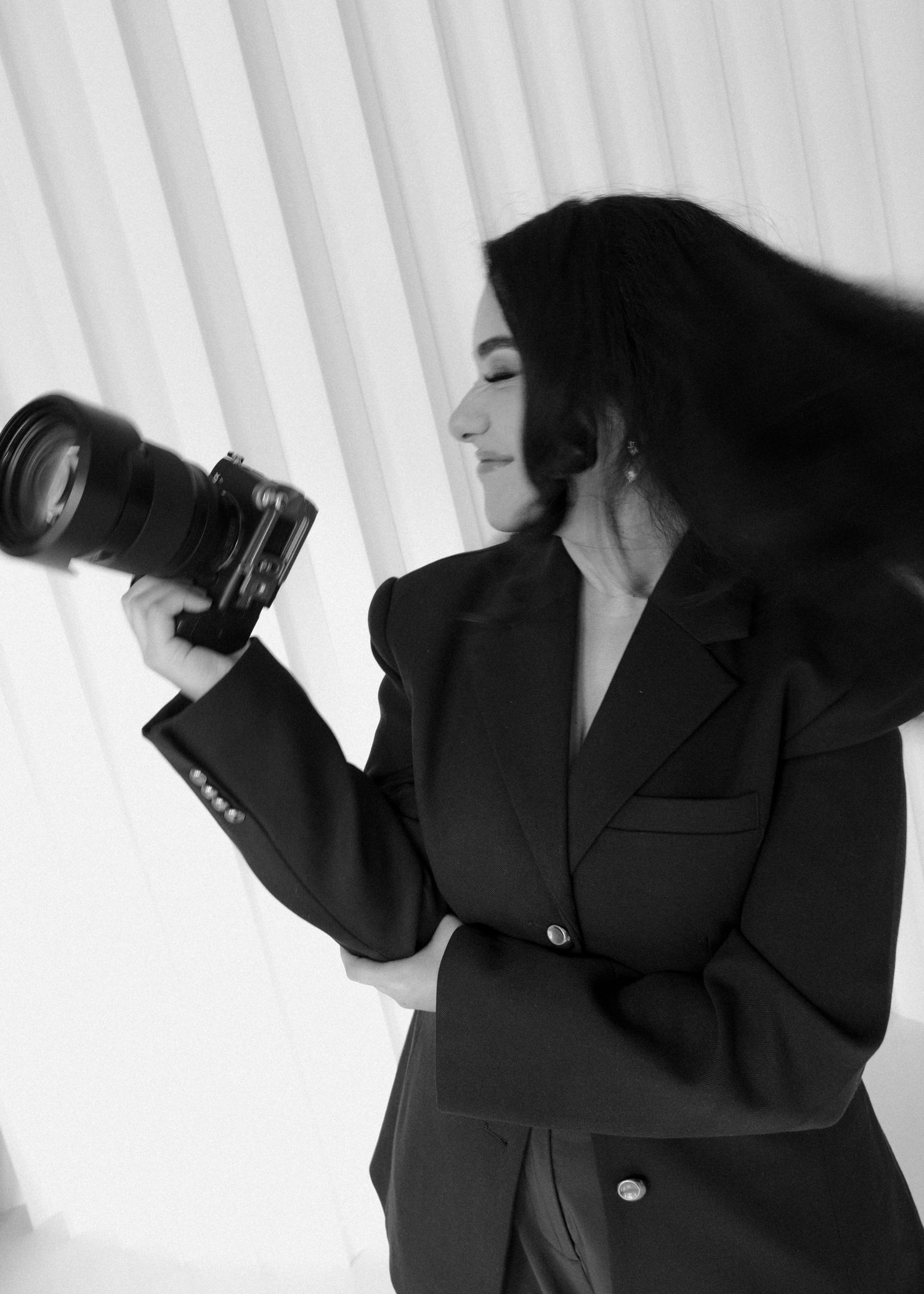 Black and white photo of a woman in a blazer holding a professional camera, with hair flowing, against a light background.