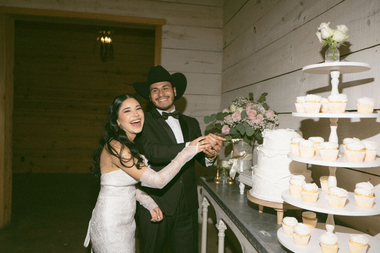 Bride and groom cutting wedding cake at rustic barn reception, groom wearing black cowboy hat and tuxedo.