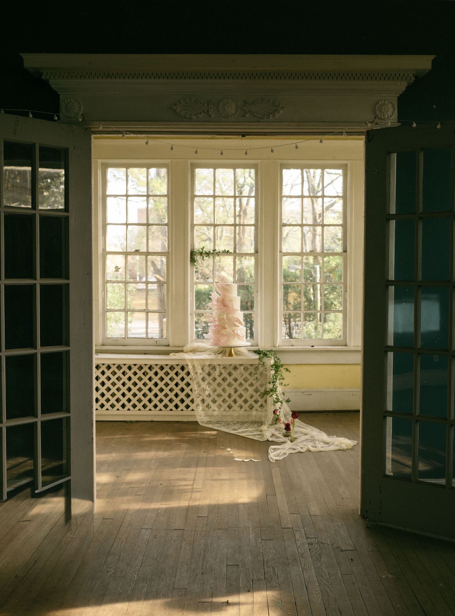 Elegant pink wedding cake displayed in sunlit room with hardwood floors and large windows in historic venue.