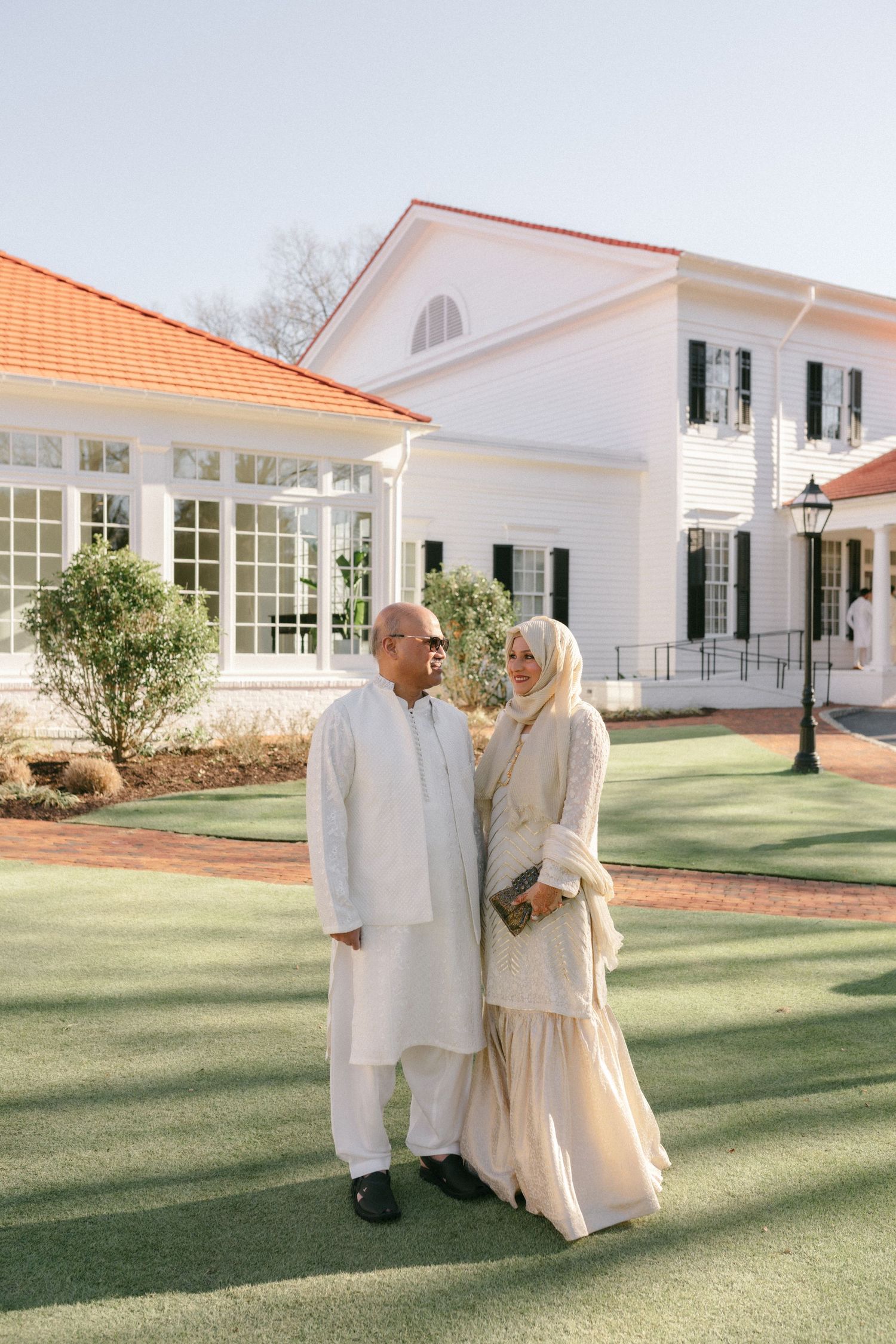 Muslim couple in cream traditional attire standing on green lawn before white colonial house with orange roof.