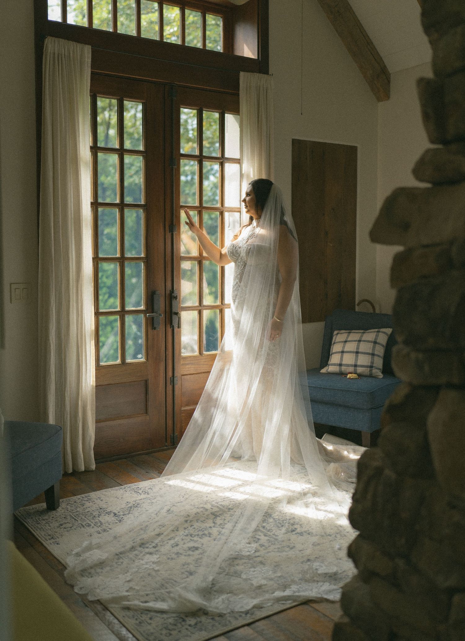 Bride in a flowing white gown and cathedral veil stands by French doors in a rustic room, bathed in natural light.