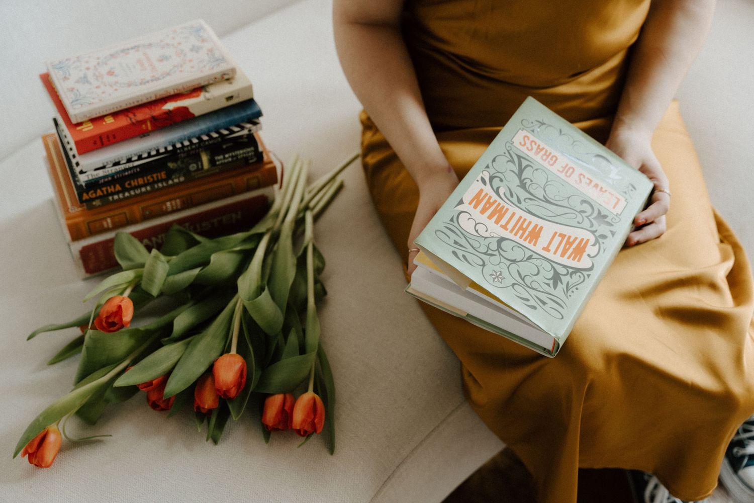 Woman in mustard dress holding a book beside stacked colorful books and fresh orange tulips on white surface.