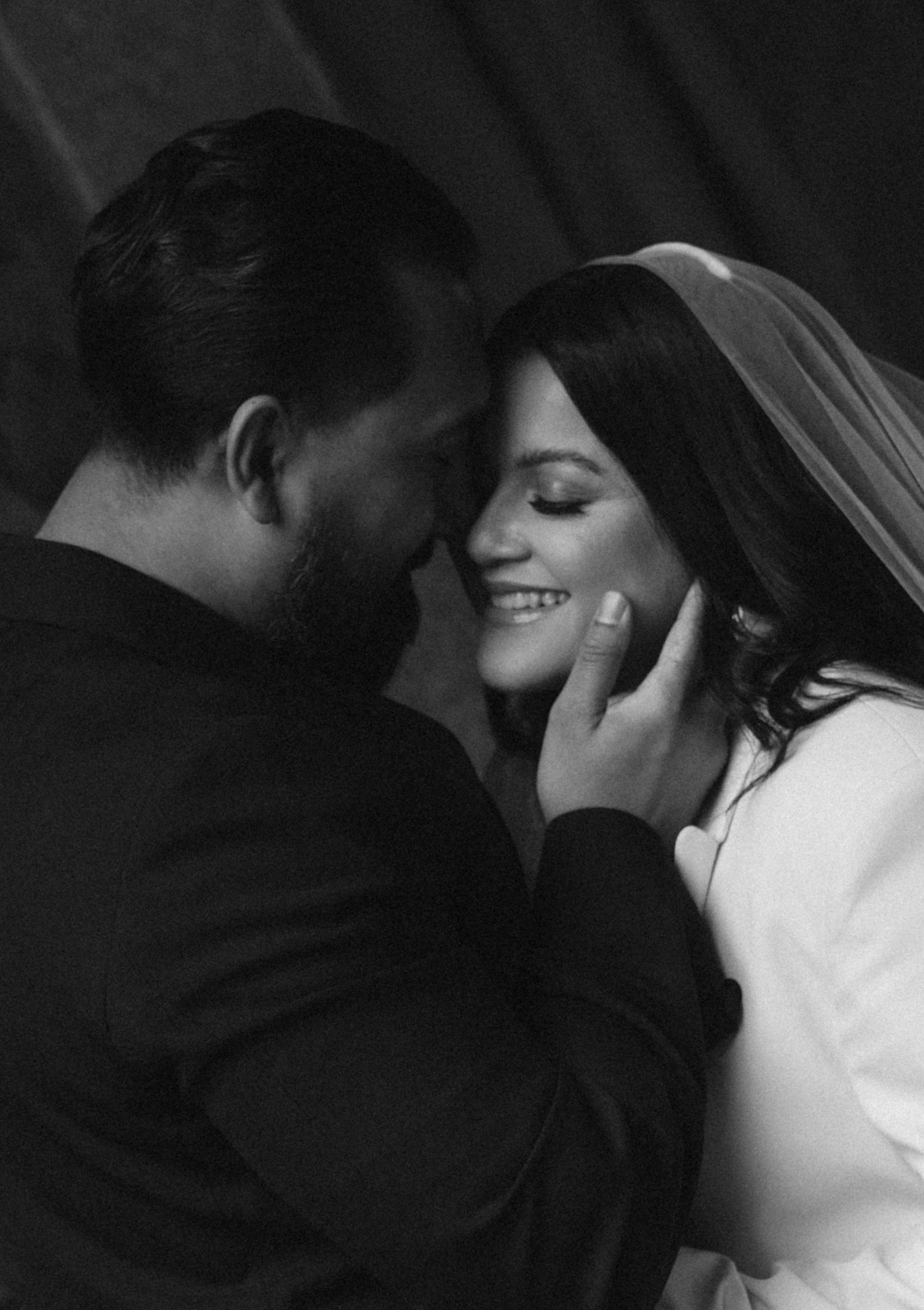 Black and white wedding photo of a smiling bride in veil and groom tenderly touching her face.