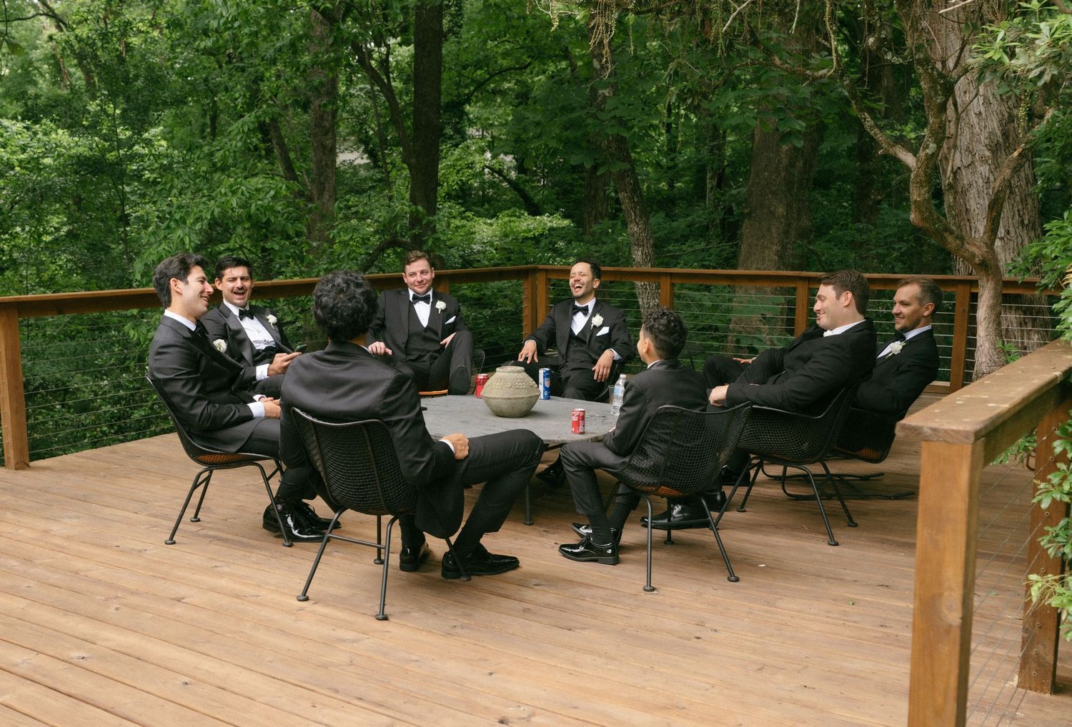 Groomsmen in black tuxedos gathered around a table on a wooden deck surrounded by lush green trees.