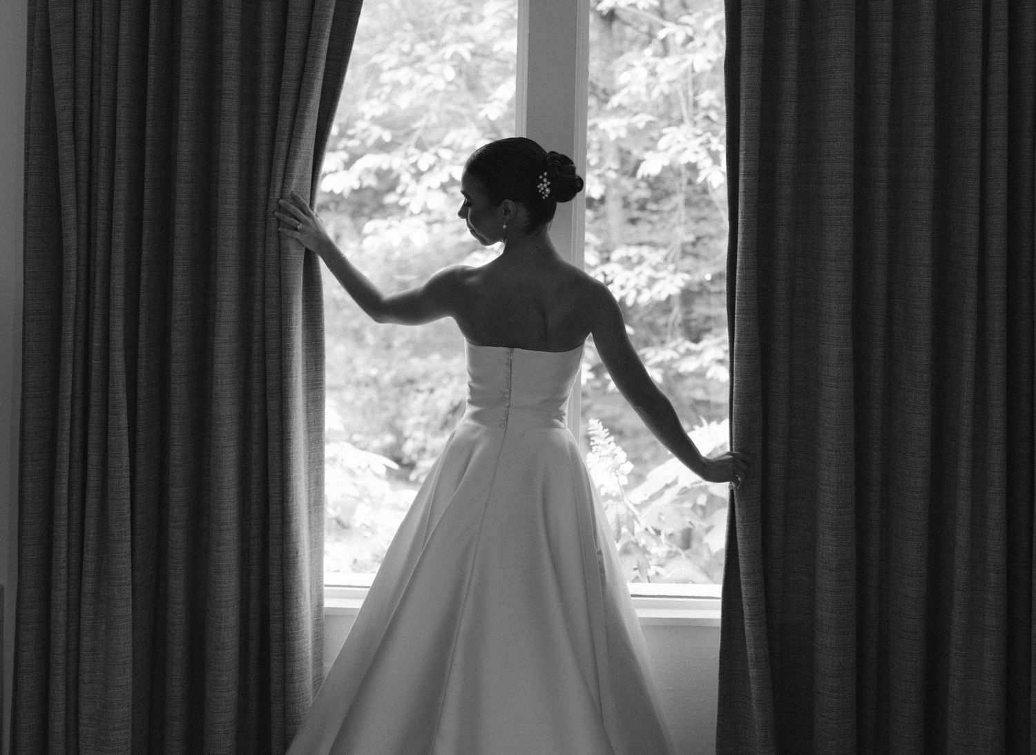 Black and white photo of a bride in a strapless wedding gown standing at a curtained window.